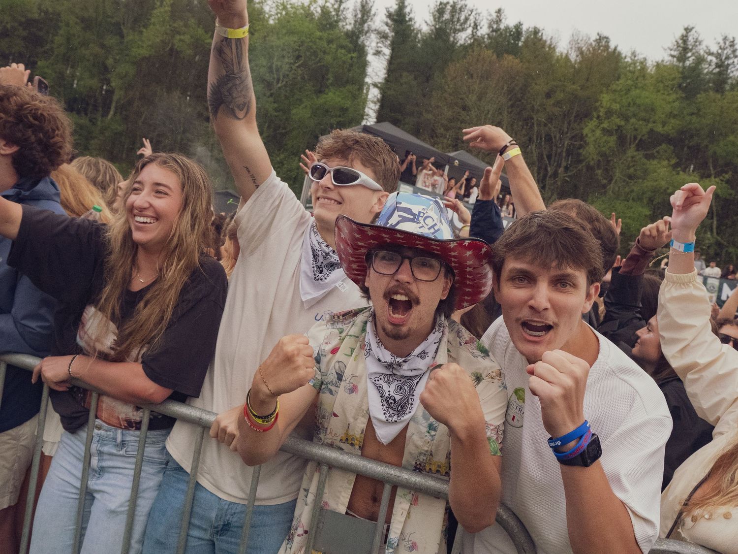 Three enthusiastic fans celebrating at outdoor music festival barrier on cloudy day.