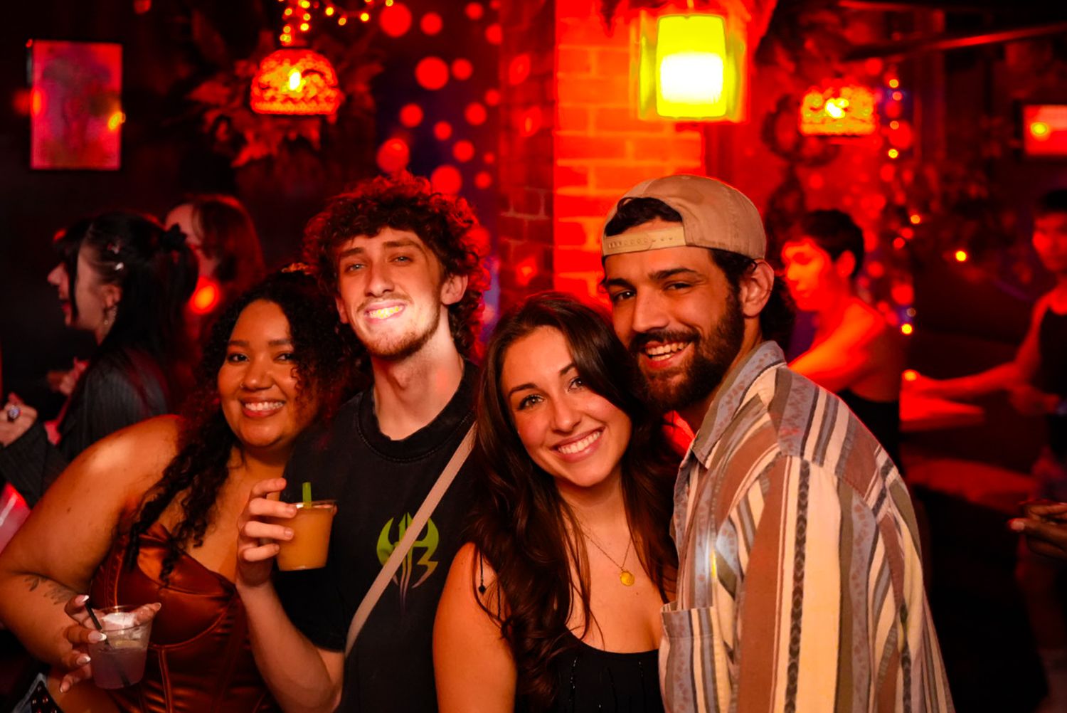 Four young friends smiling and posing together at a vibrant red-lit nightclub party with drinks.