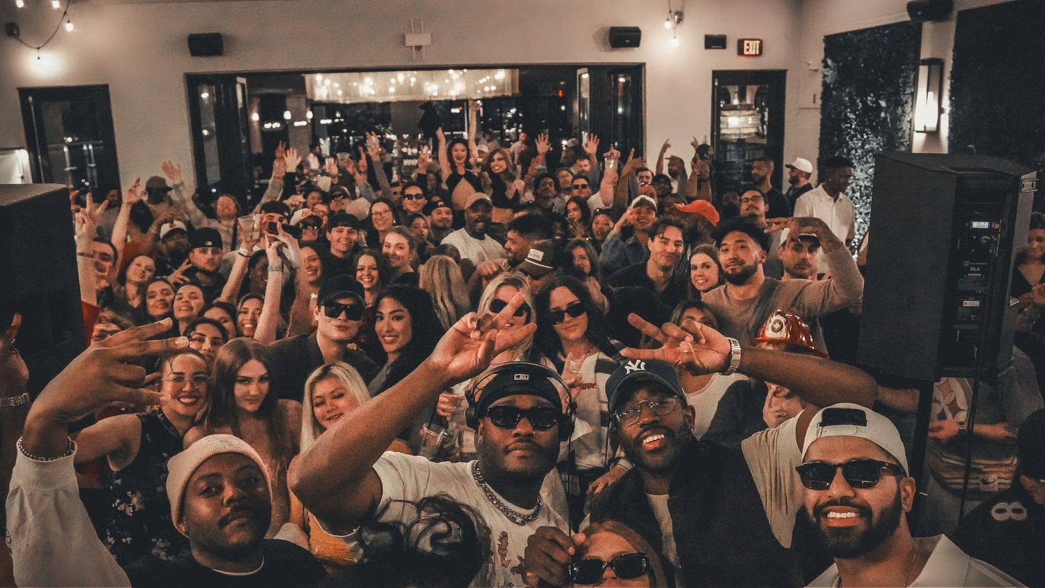 Large diverse crowd at an indoor party event, raising hands and taking a group selfie with DJ and performers.