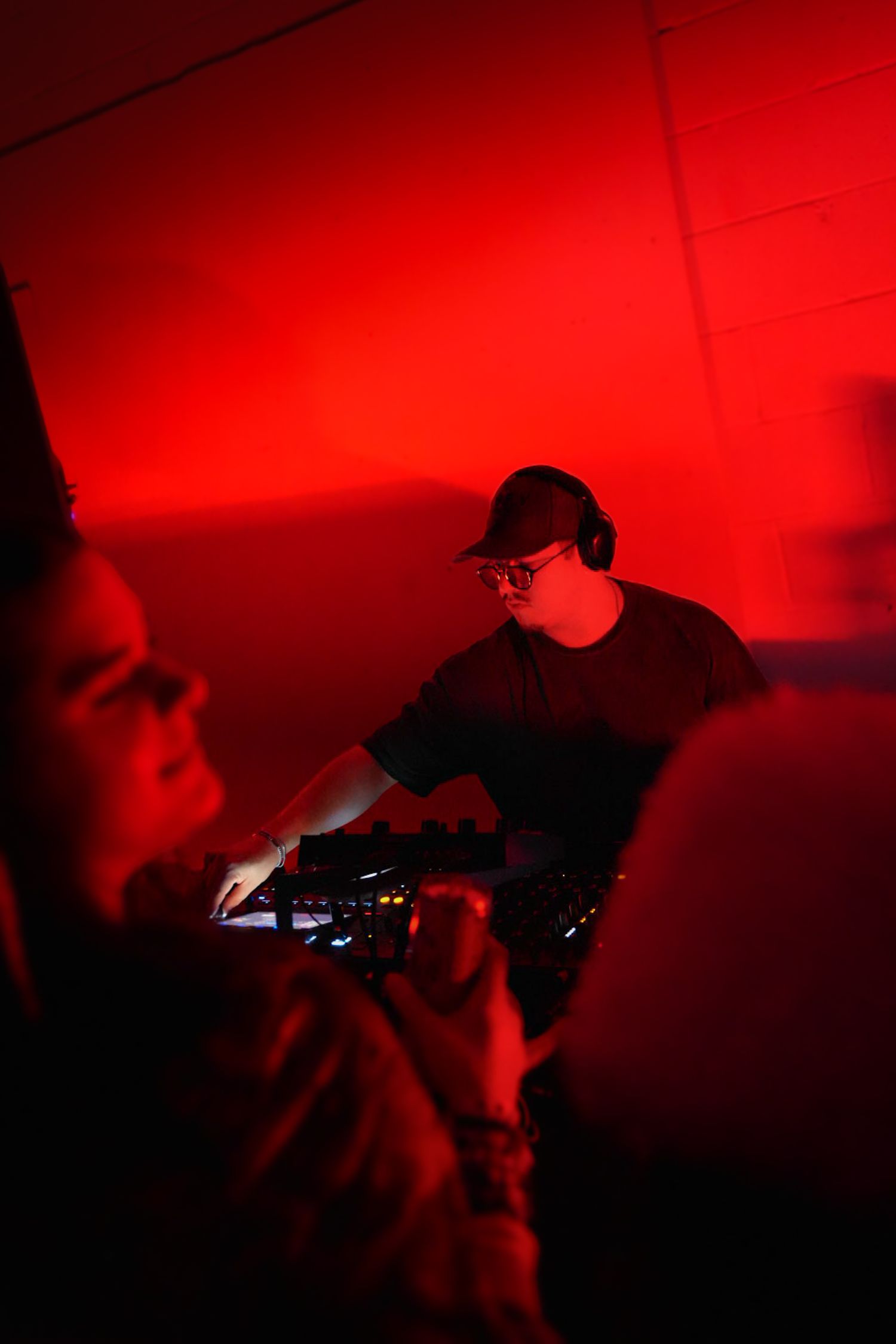 DJ wearing glasses and headphones mixing tracks at a red-lit underground club event.