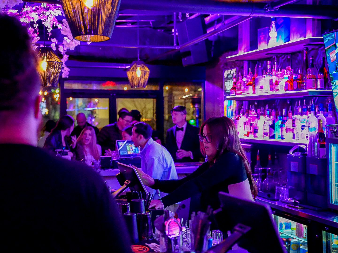 Vibrant nightclub bar scene with colorful neon lights, bartenders serving drinks, and shelves of liquor bottles.