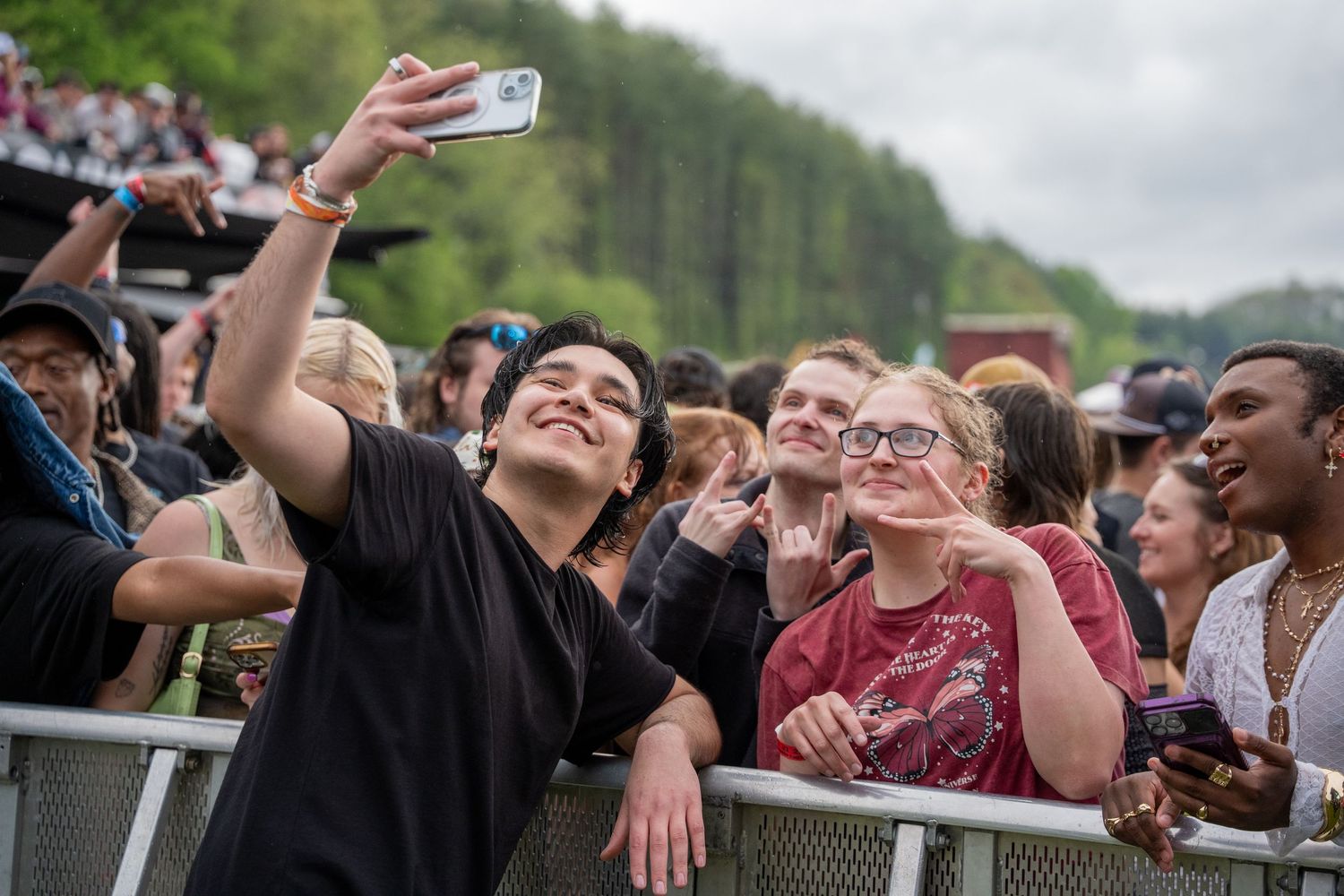 Artist taking selfie with smiling fans at outdoor music festival barrier on cloudy day.