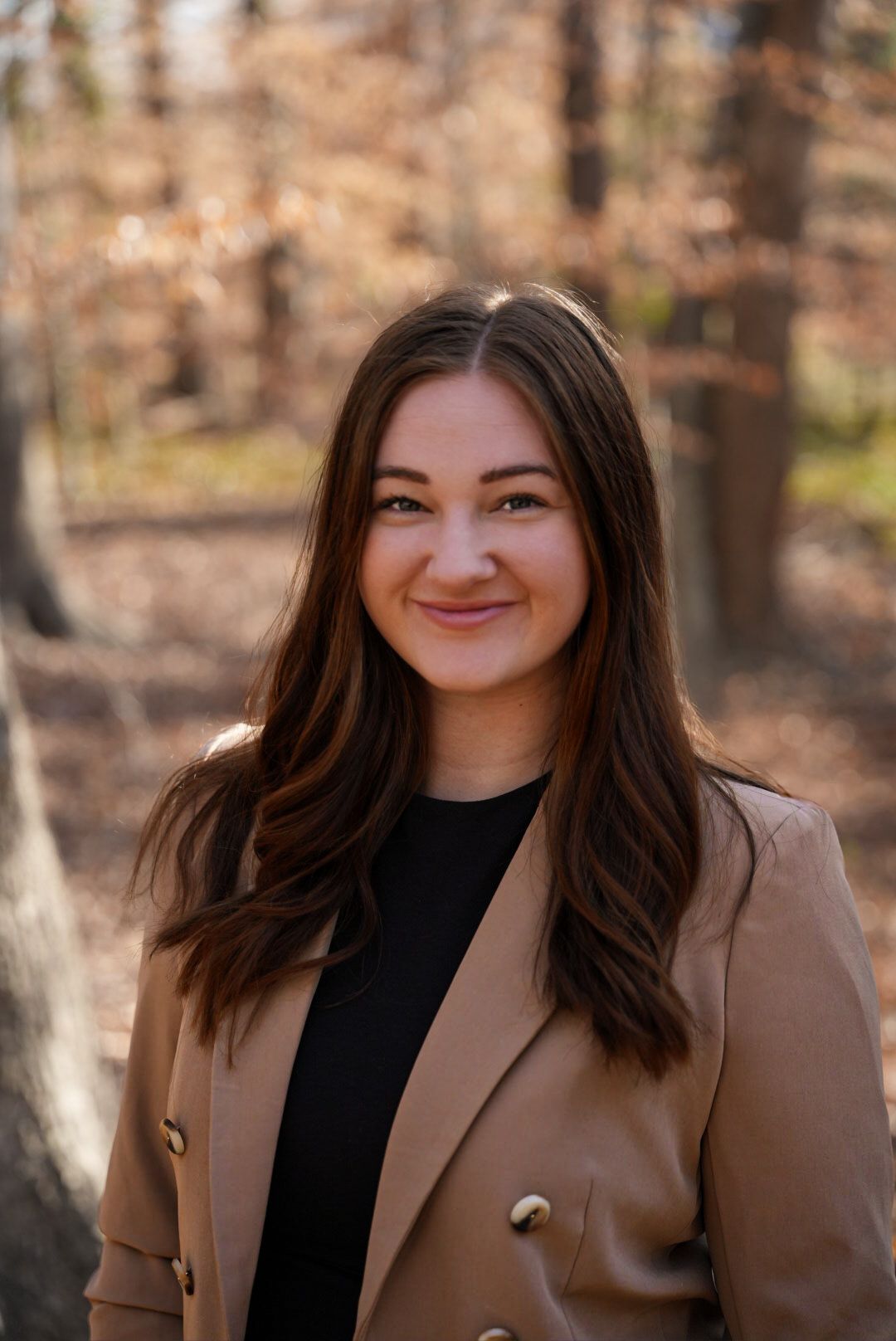 Smiling young woman with long brown hair wearing a tan blazer outdoors in an autumn forest setting.