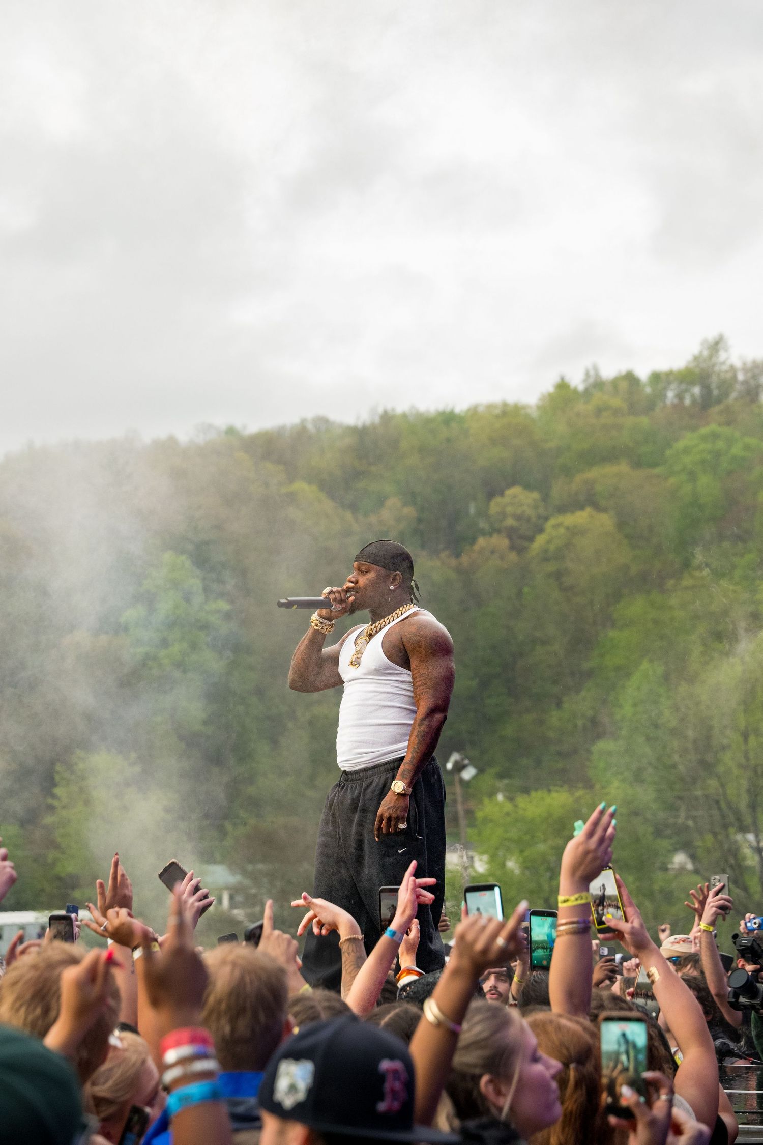 Male rapper in white tank top performs with microphone above festival crowd outdoors.