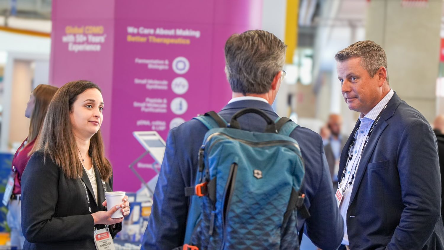 Three professionals networking at a conference booth with purple banner in exhibition hall.