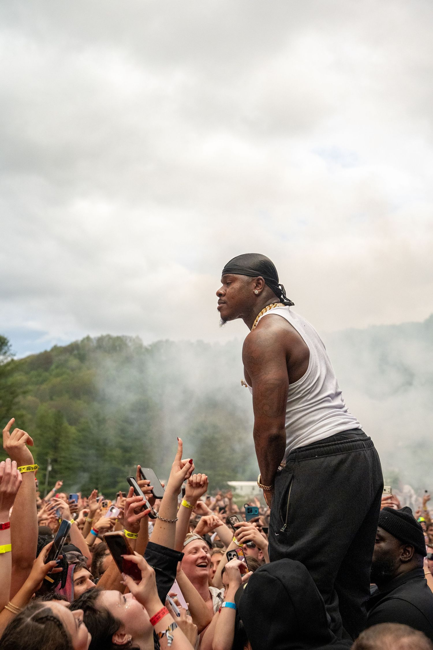 Tall male performer in white tank top stands above cheering crowd at smoky outdoor music festival.