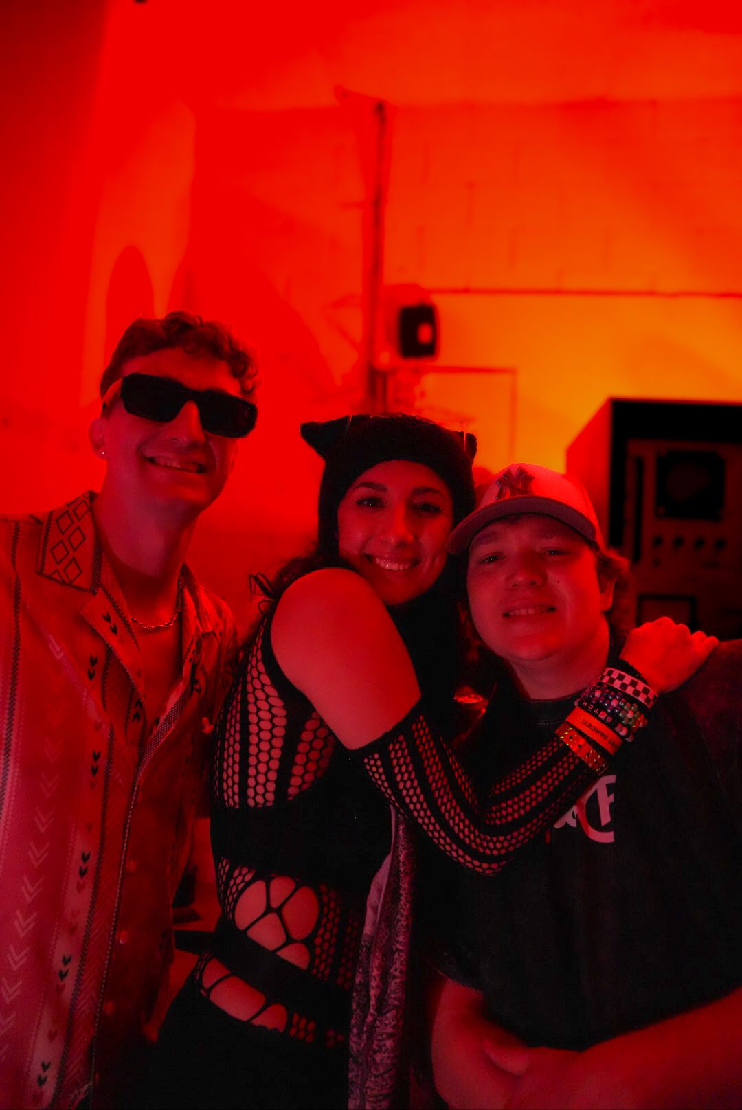 Three partygoers smile together at a red-lit nightclub event, wearing festive costumes.