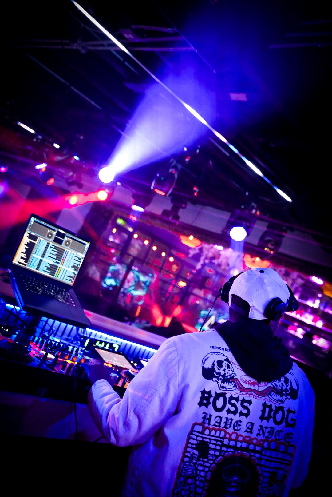 DJ in white Boss Dog hoodie and cap mixing music at a nightclub with purple laser lights.