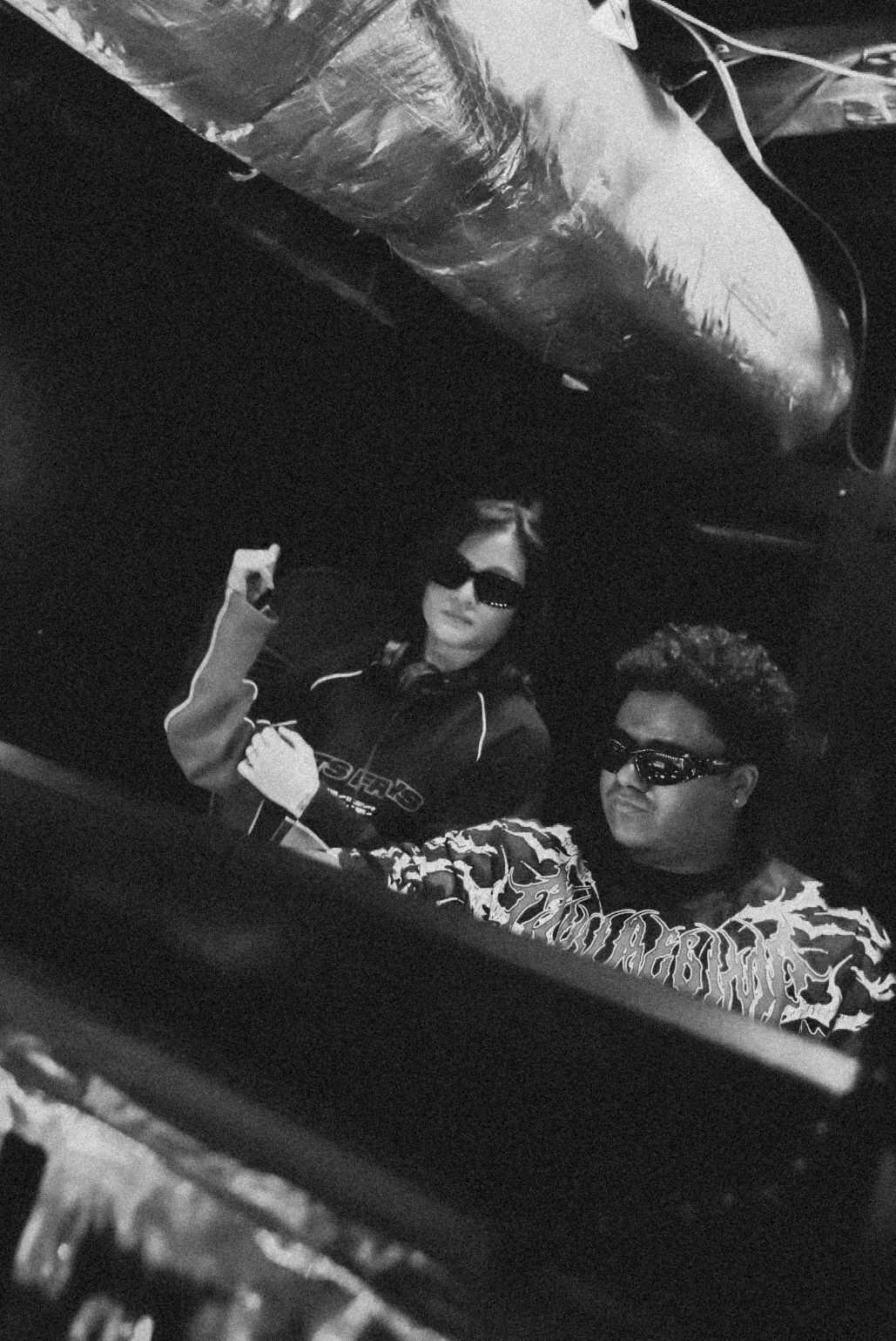 Black and white photo of two DJs working together behind equipment at a nightclub under ductwork.