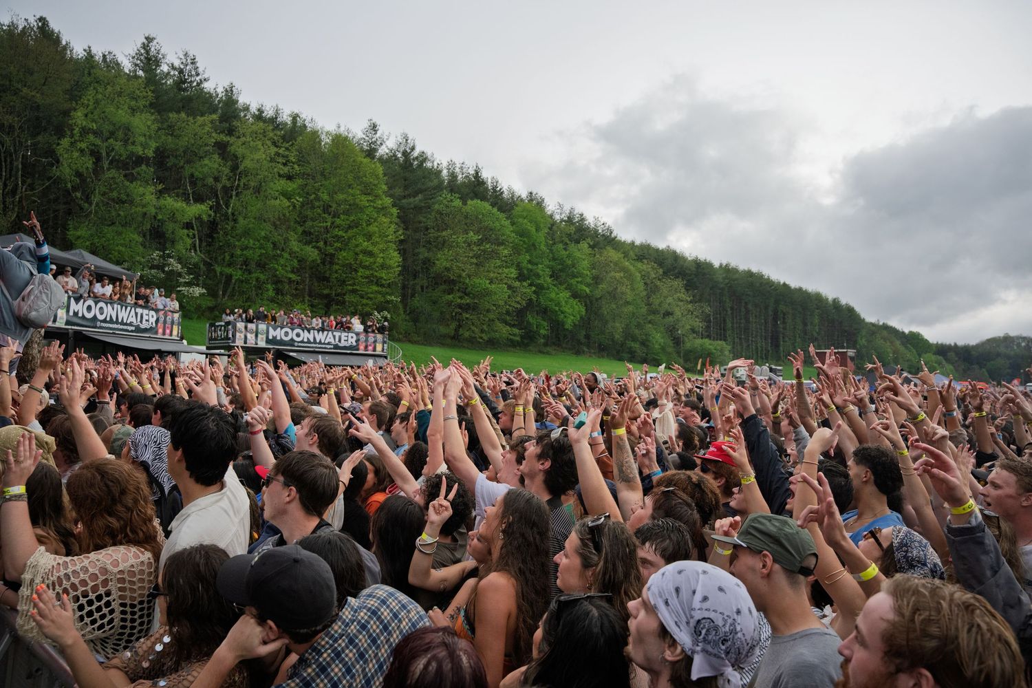 Massive crowd with raised hands at Moonwater outdoor music festival surrounded by forest.
