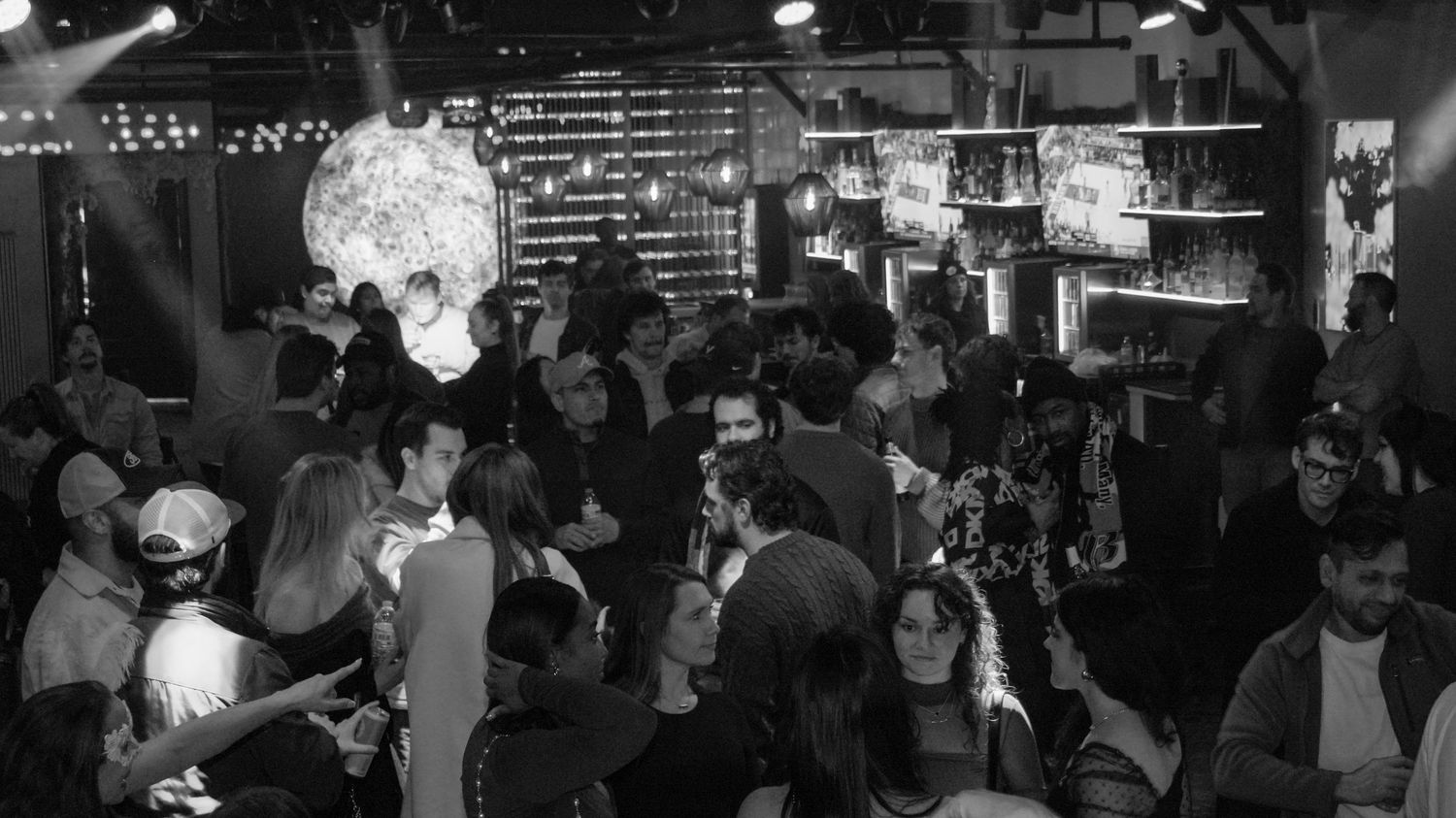 Black and white wide-angle shot of a crowded nightclub dance floor with a disco ball and bar area.