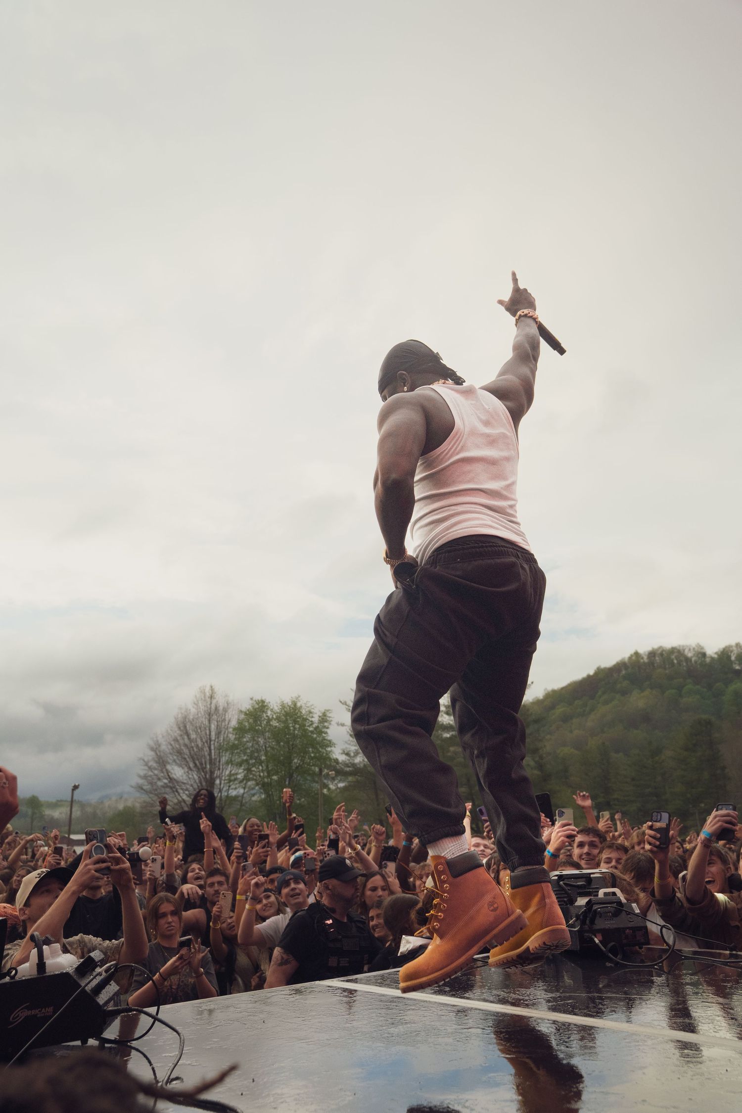 Rapper performing on stage with mic raised, wearing white tank top and Timberland boots, energizing a large outdoor festival crowd.
