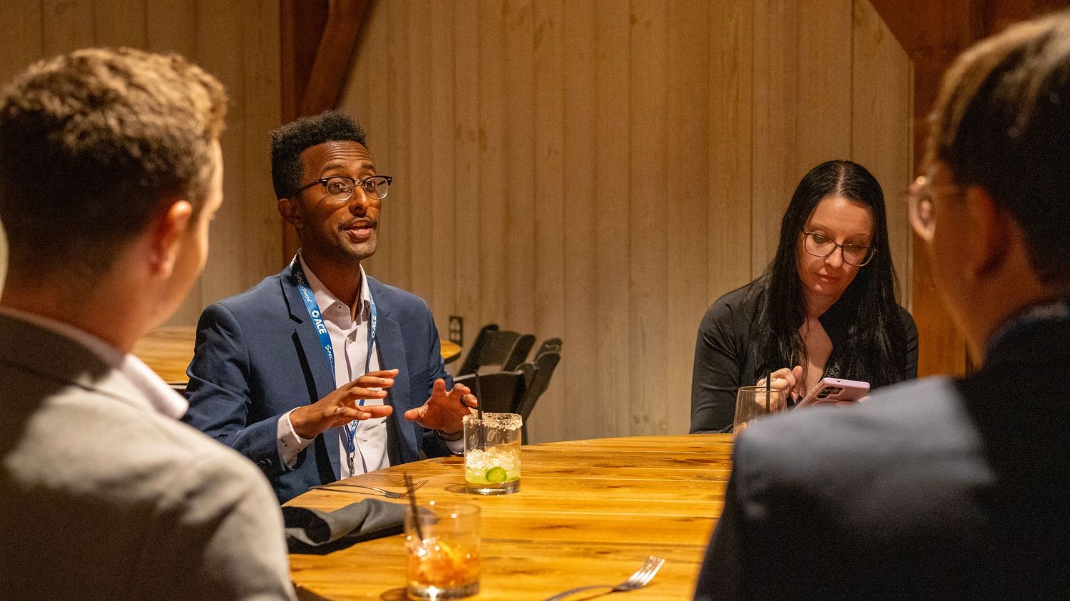Business professionals engaged in discussion around wooden table in warmly lit restaurant setting.