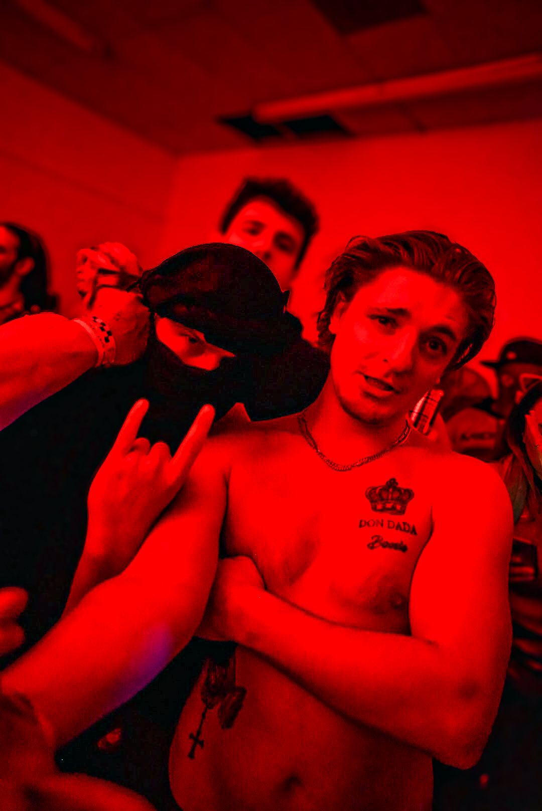Shirtless young man with chest tattoo poses with friends under red club lighting.