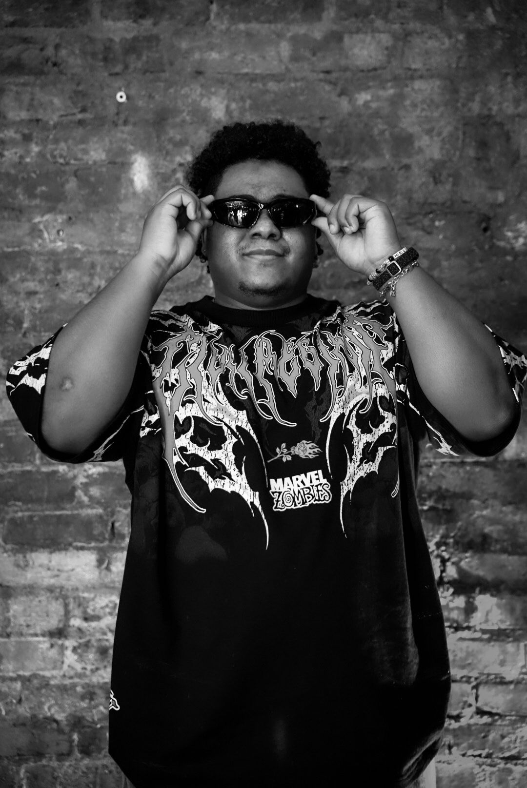 Smiling young man adjusting sunglasses in black and white portrait against brick wall.