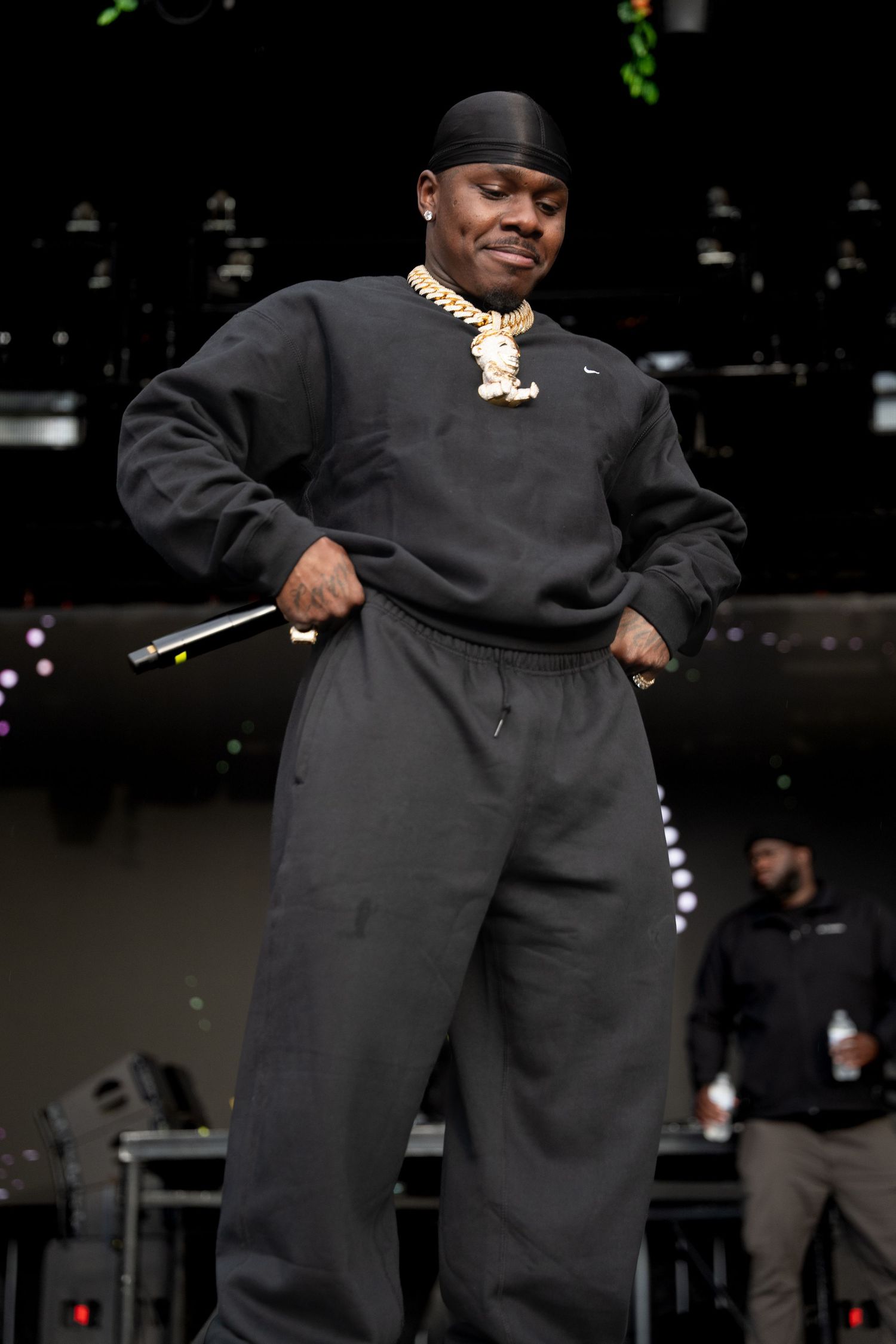 Rapper in black sweatsuit holding microphone poses confidently on outdoor festival stage with floral garland decorations behind him.
