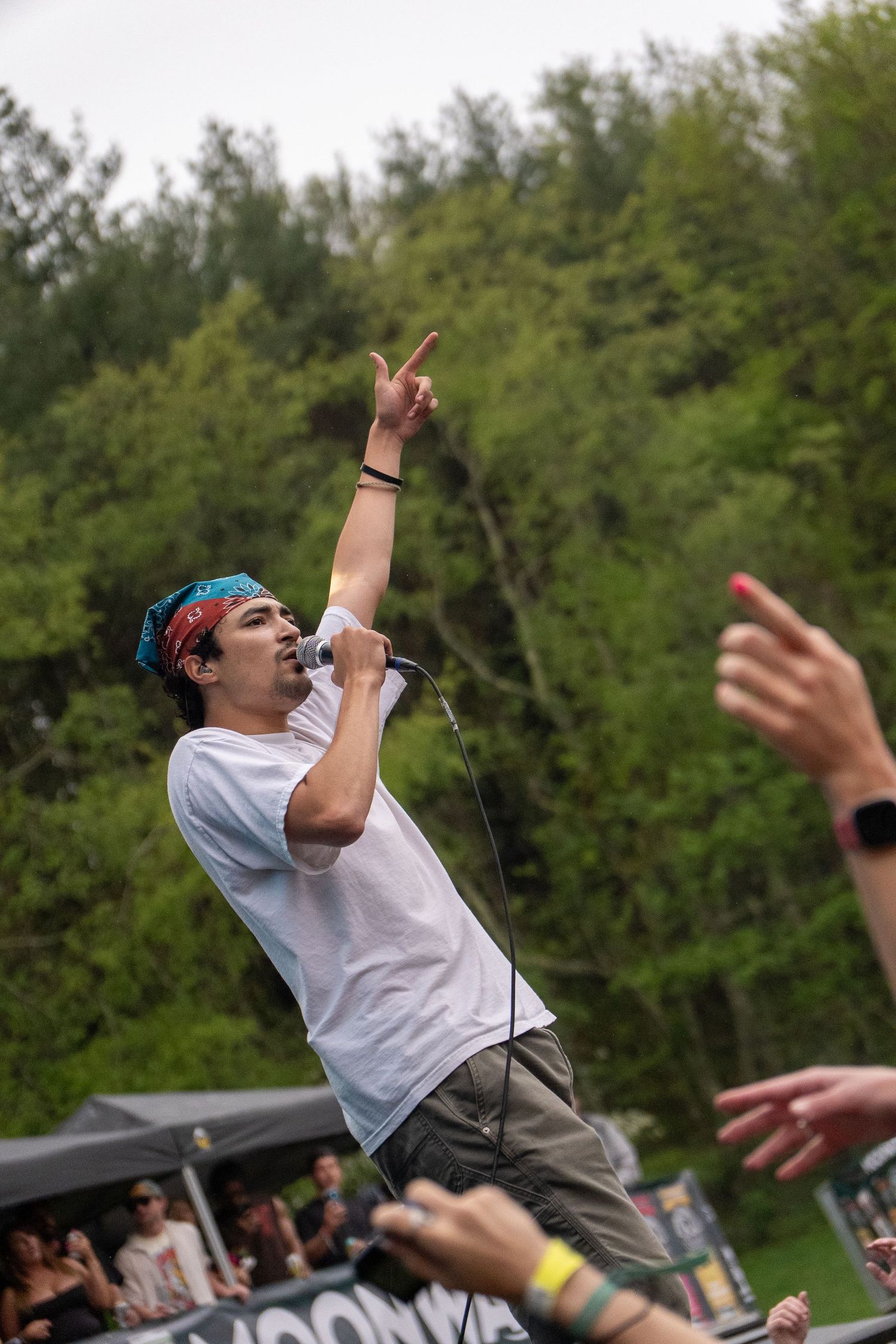 Energetic male rapper pointing skyward while performing at outdoor music festival with crowd reaching toward stage.