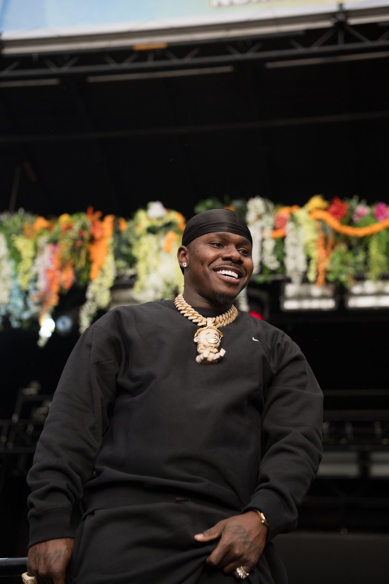 Smiling rapper in black Nike sweatshirt and gold chain necklace sits relaxed in front of floral-decorated festival stage.
