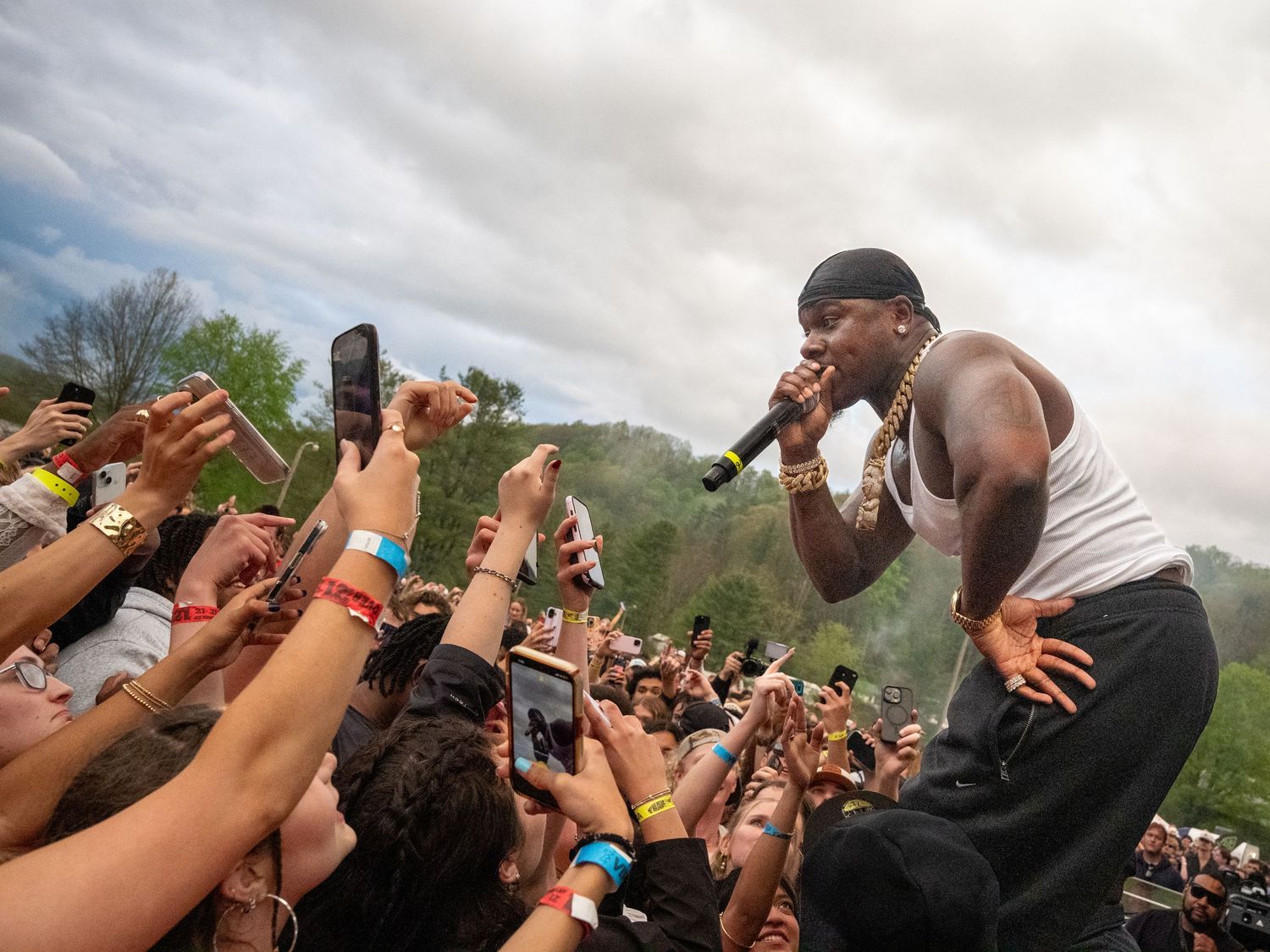 Muscular male rapper in white tank and gold chain performs energetically for massive outdoor festival crowd.