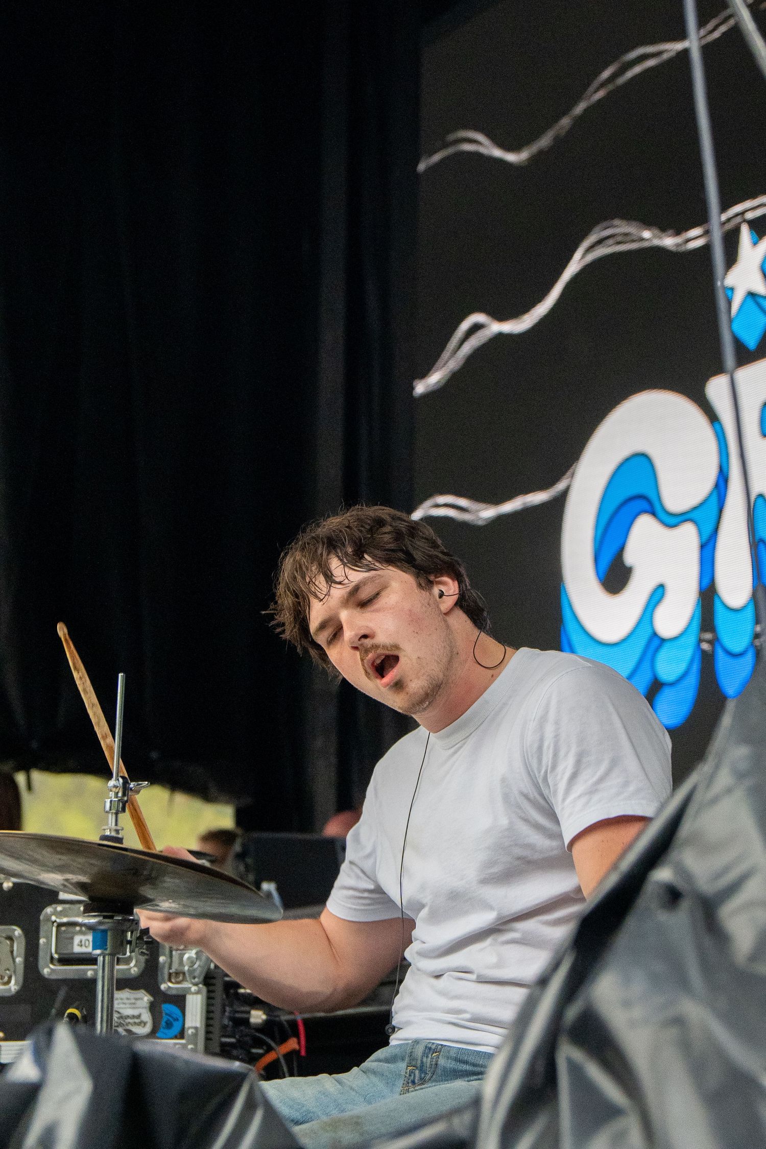 Passionate drummer performing on stage with band logo backdrop at outdoor music festival.