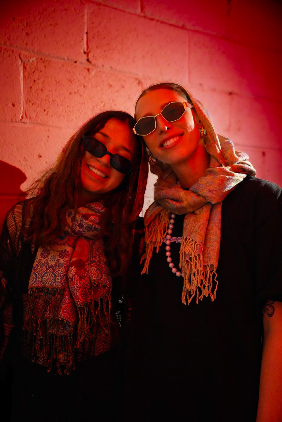 Two smiling women wearing sunglasses and scarves posing against red-lit brick wall.