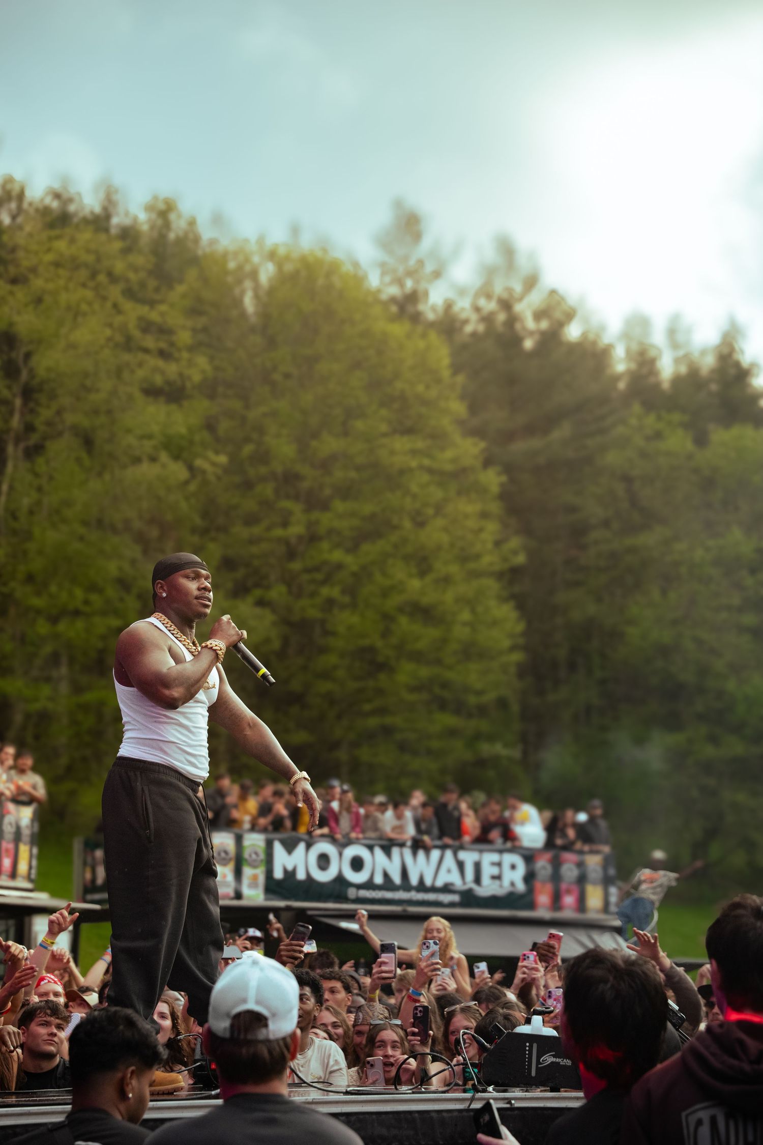 Rapper in white tank top performs on outdoor stage at Moonwater festival surrounded by dense crowd and lush green trees.