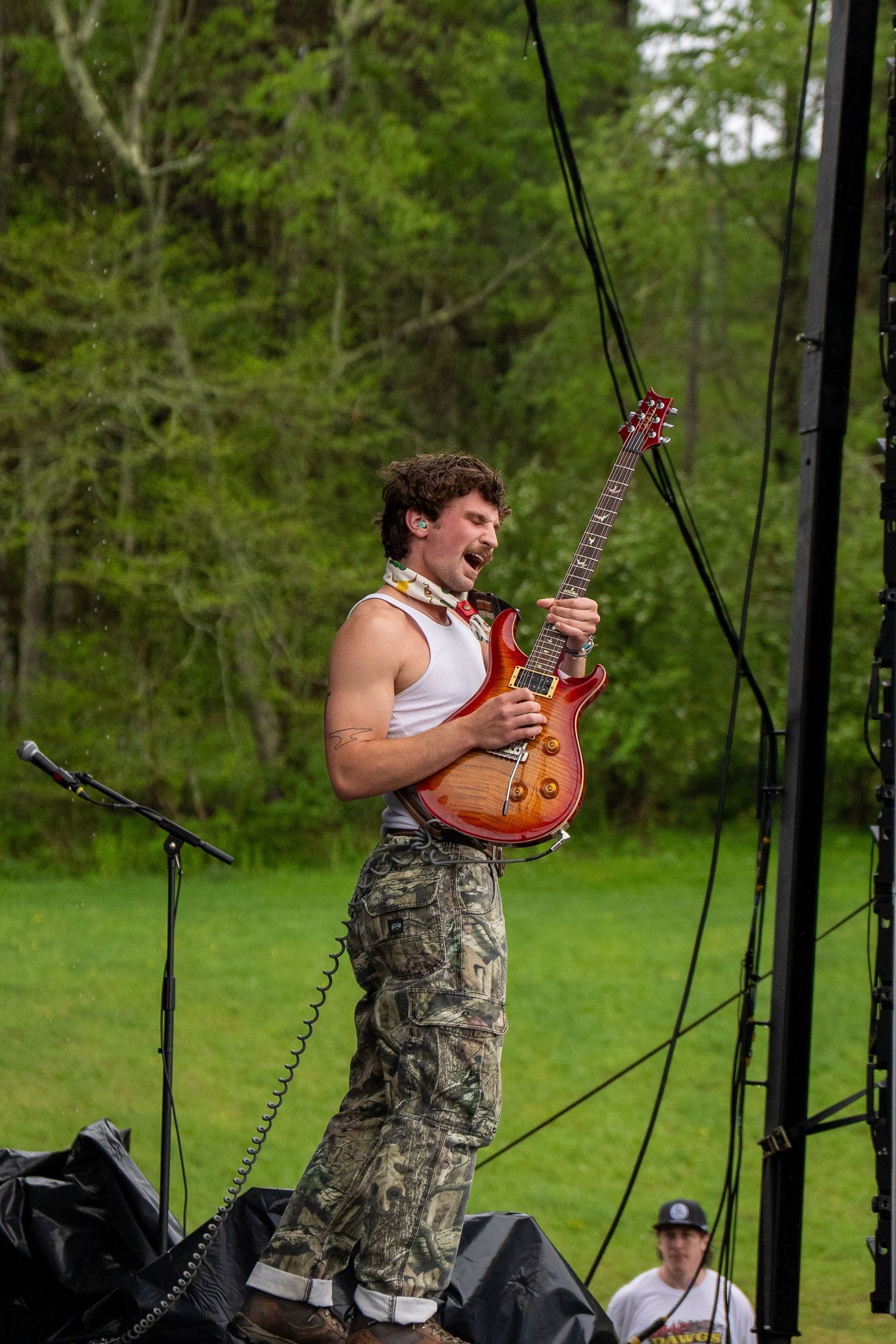 Shirtless guitarist in camo pants performs passionately on outdoor festival stage holding red electric guitar.
