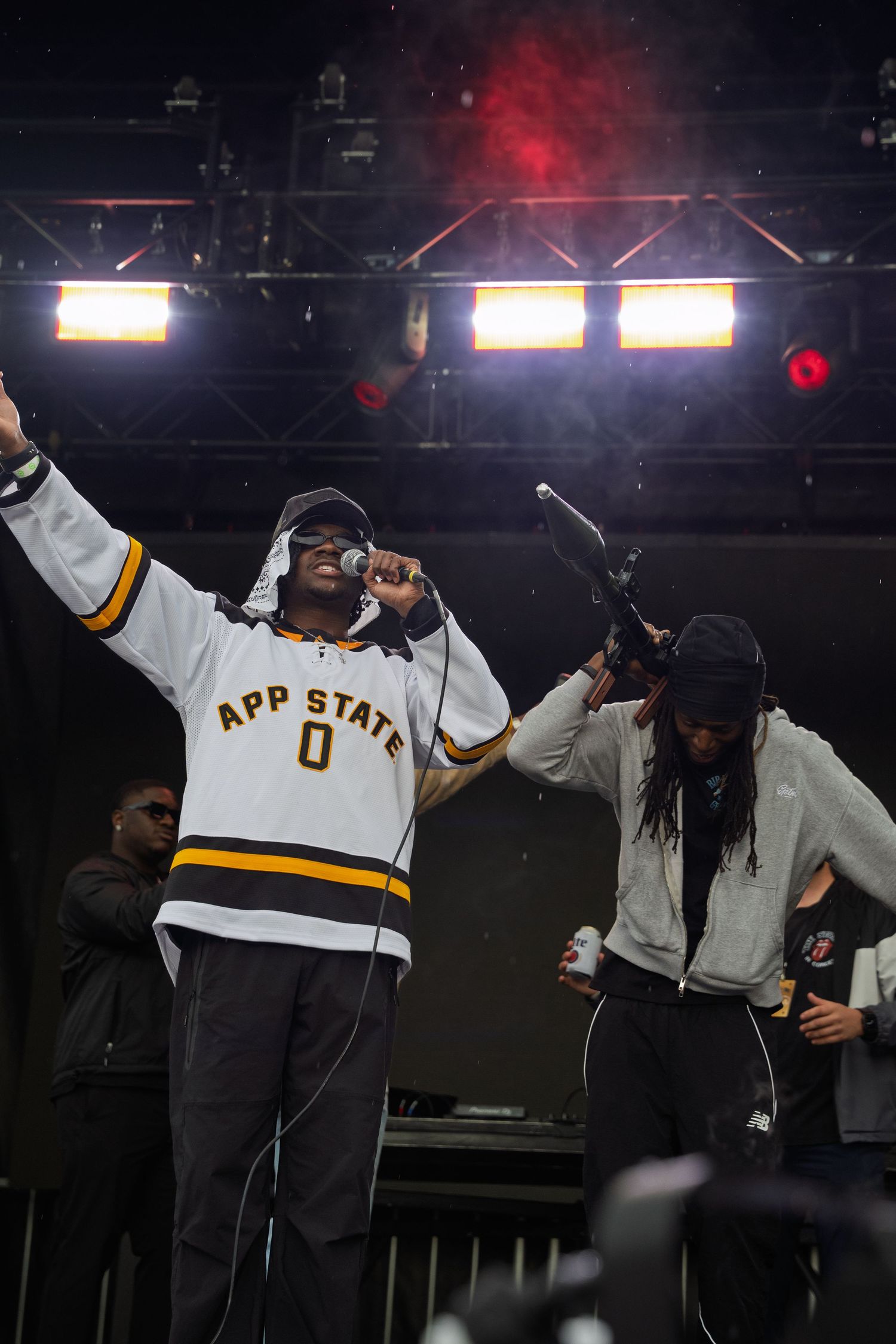 Performer in App State jersey raises arm on stage with bandmate holding bottle at festival.