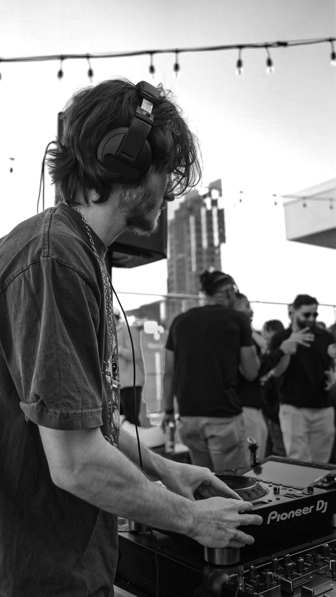 A DJ wearing headphones operates a Pioneer DJ mixer at an outdoor rooftop party.