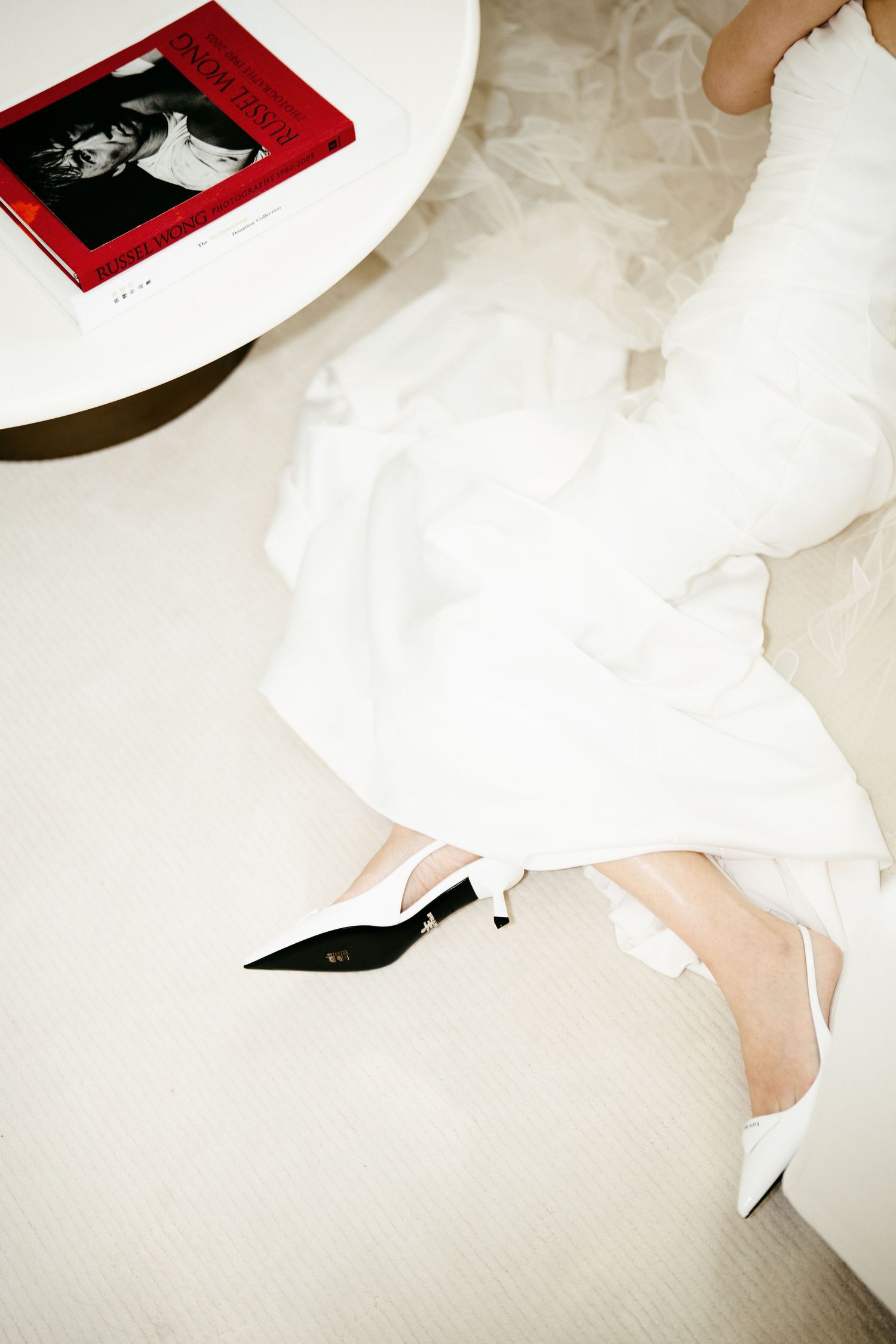Bride wearing black and white slingback heels with flowing white wedding dress beside a red book on a table.