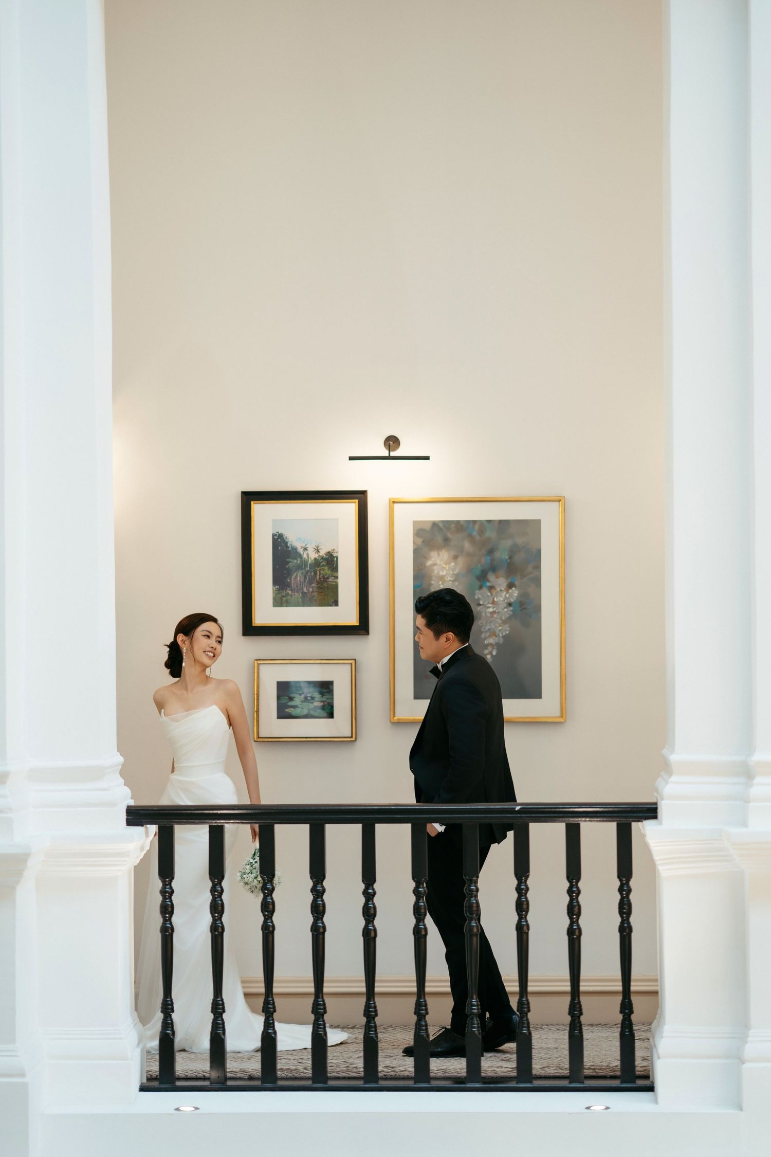 Couple stands at a white balcony with framed artwork on the wall during an elegant wedding photoshoot.