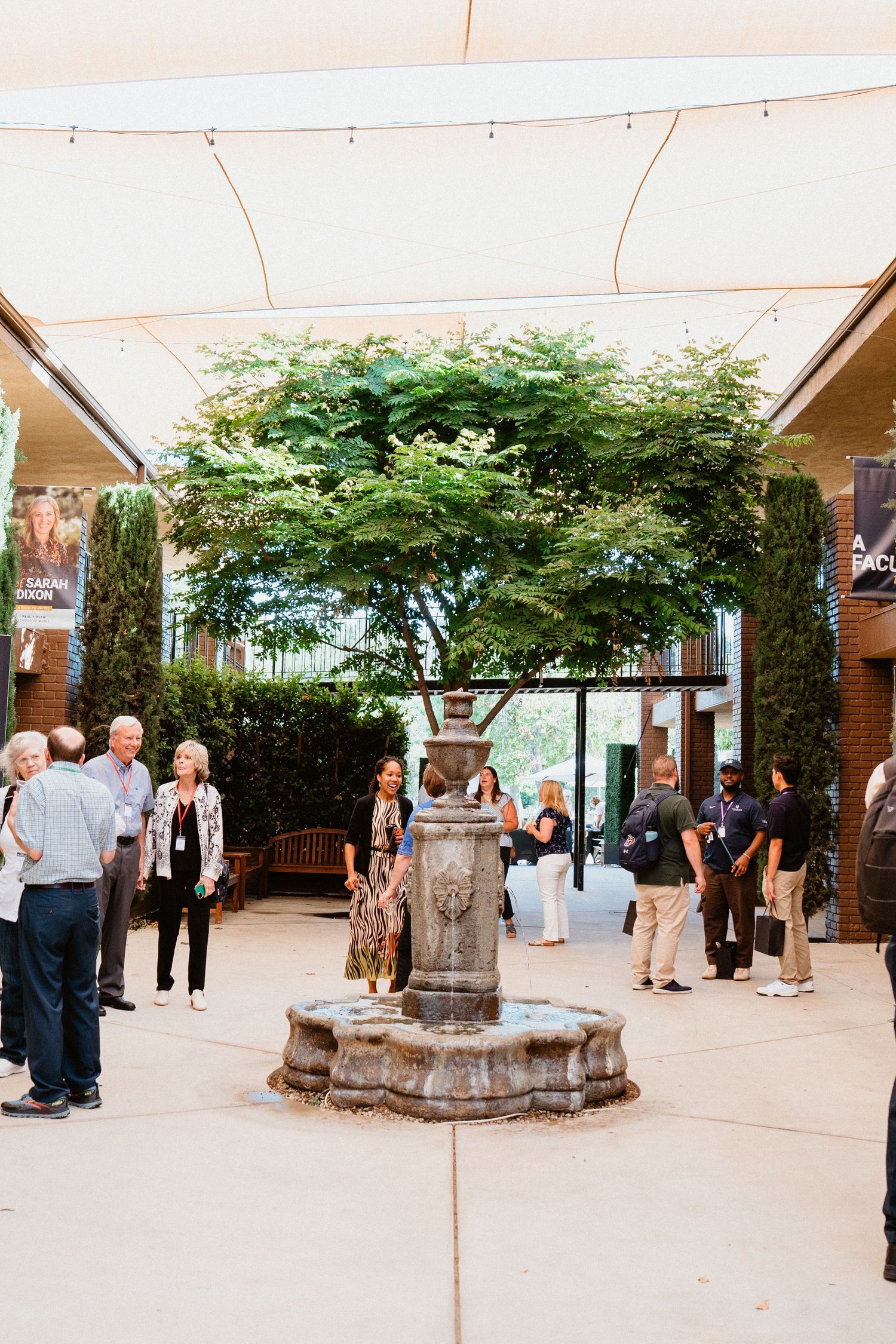 People gather around a decorative stone fountain in an outdoor courtyard shaded by a large green tree.