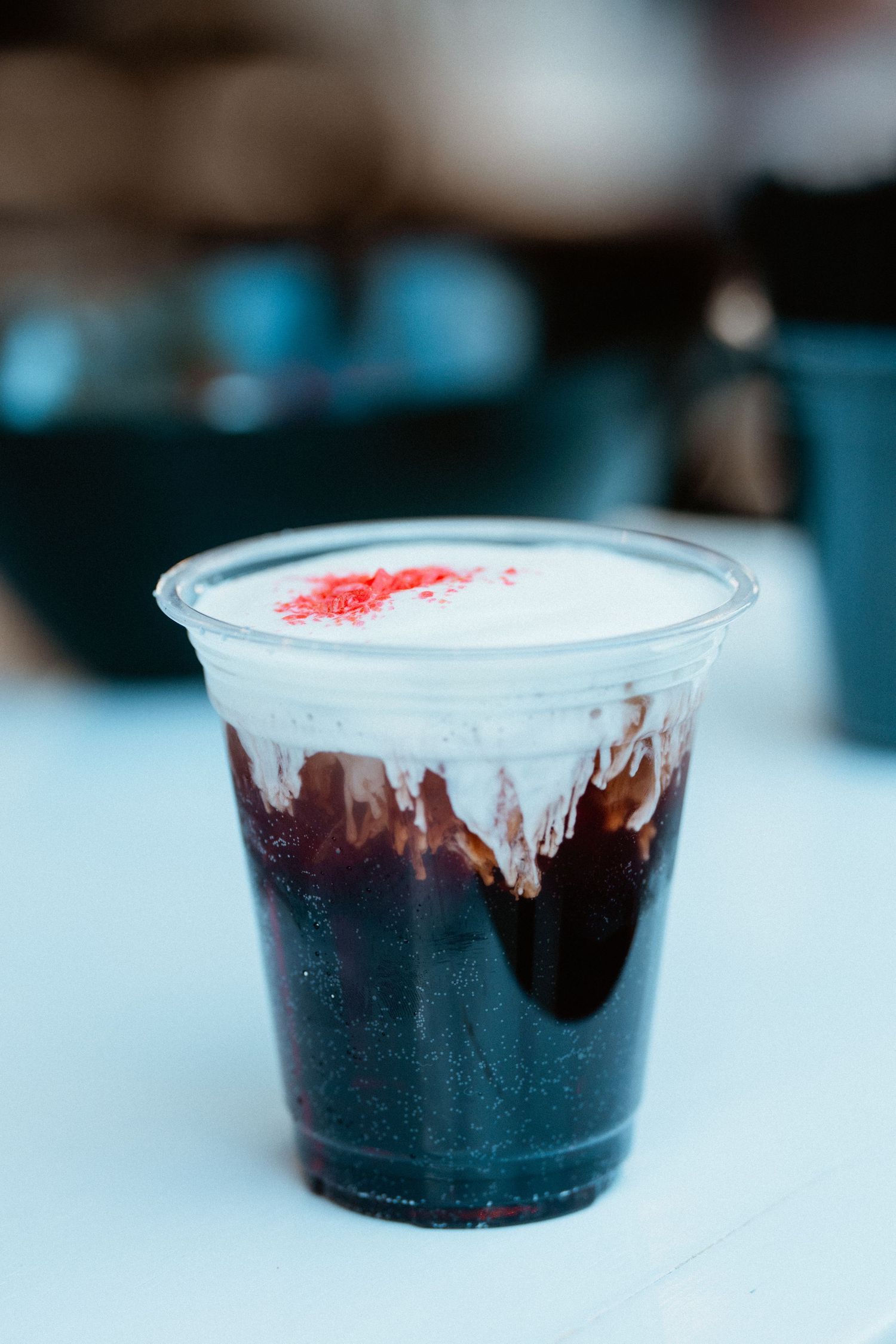 A dark beverage in a clear plastic cup topped with whipped cream and red sprinkles on a white surface.