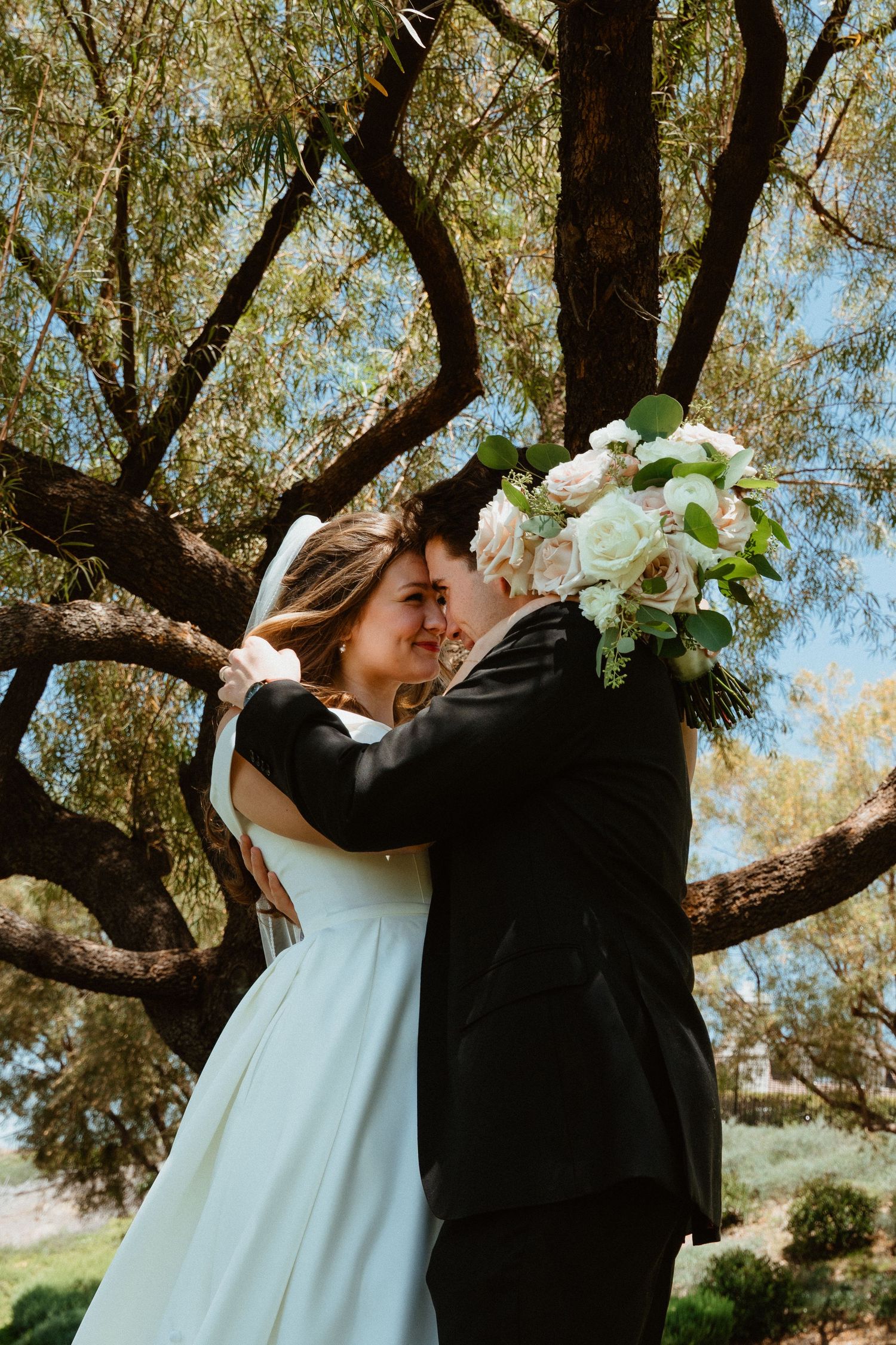 Bride and groom embrace and smile under a large tree outdoors, bride holding a white and blush rose bouquet.