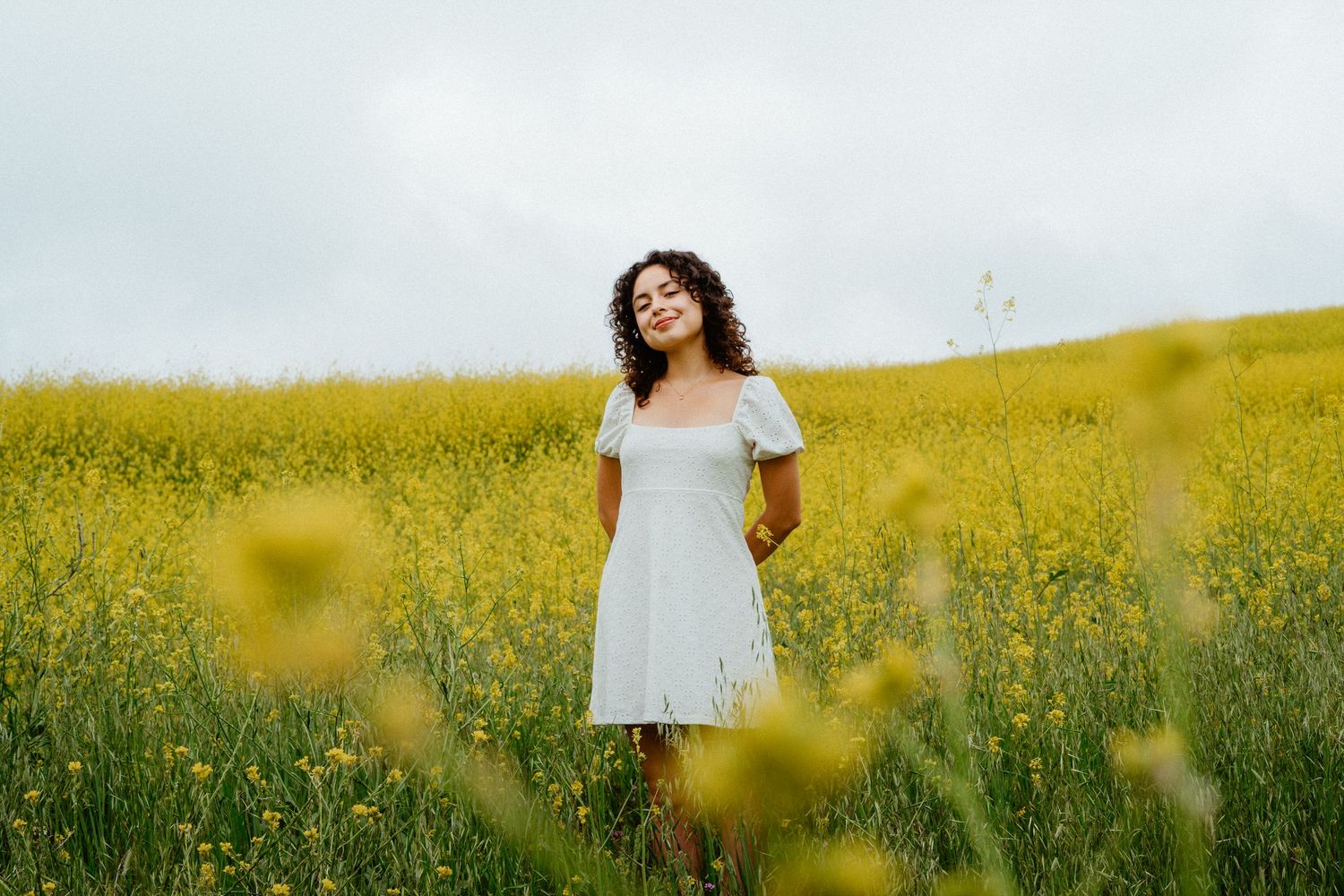 Young woman with curly hair in white dress standing in a vibrant yellow flower field.