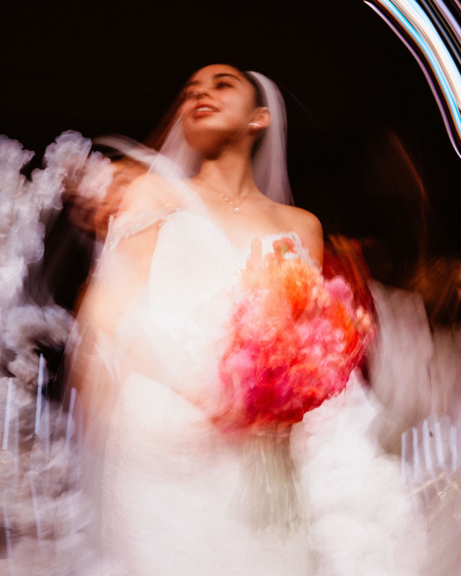 Dreamy motion blur portrait of smiling bride in white gown and veil holding vibrant pink and orange floral bouquet.