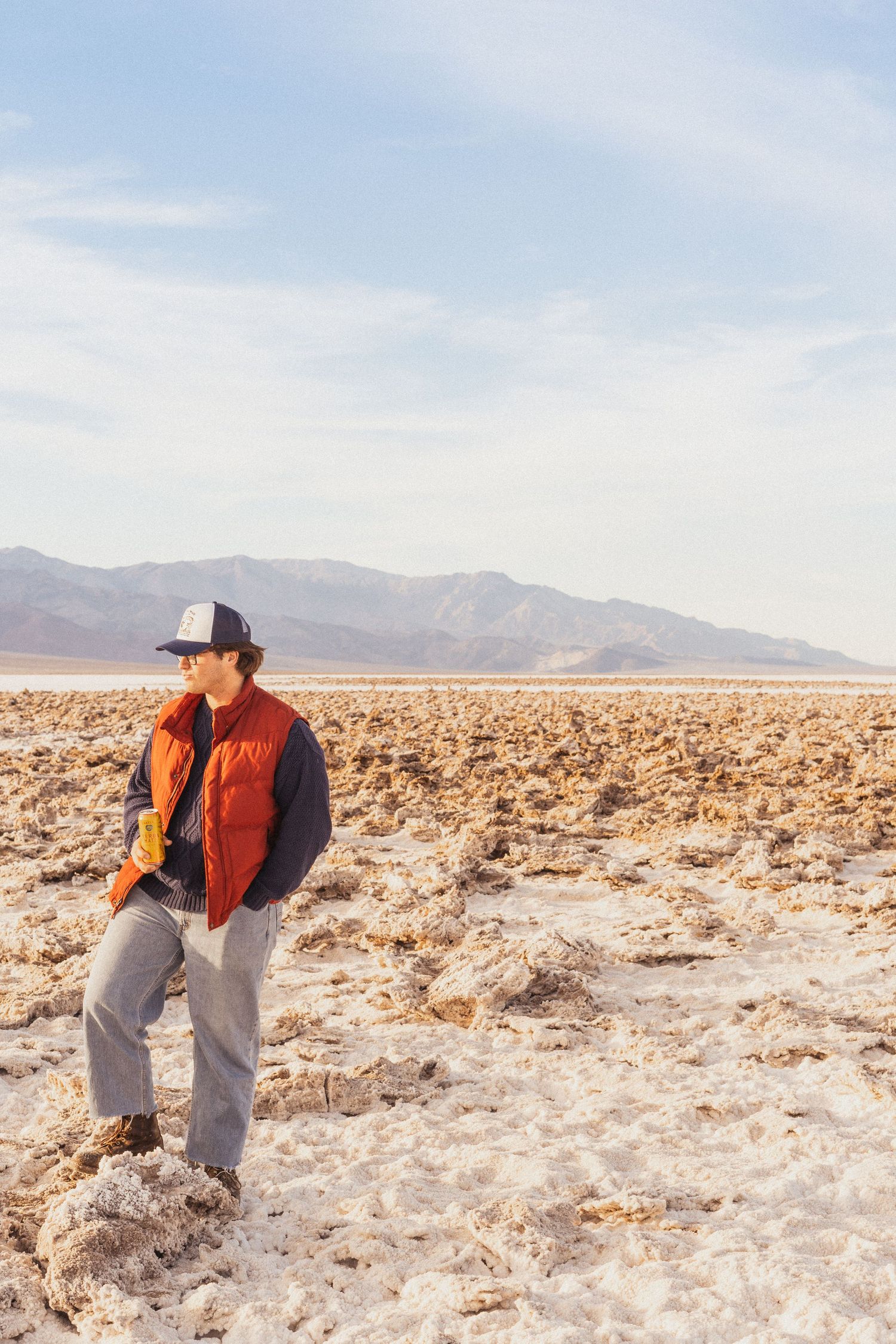 A person in an orange vest and trucker hat stands on a white salt flat with mountains in the background.