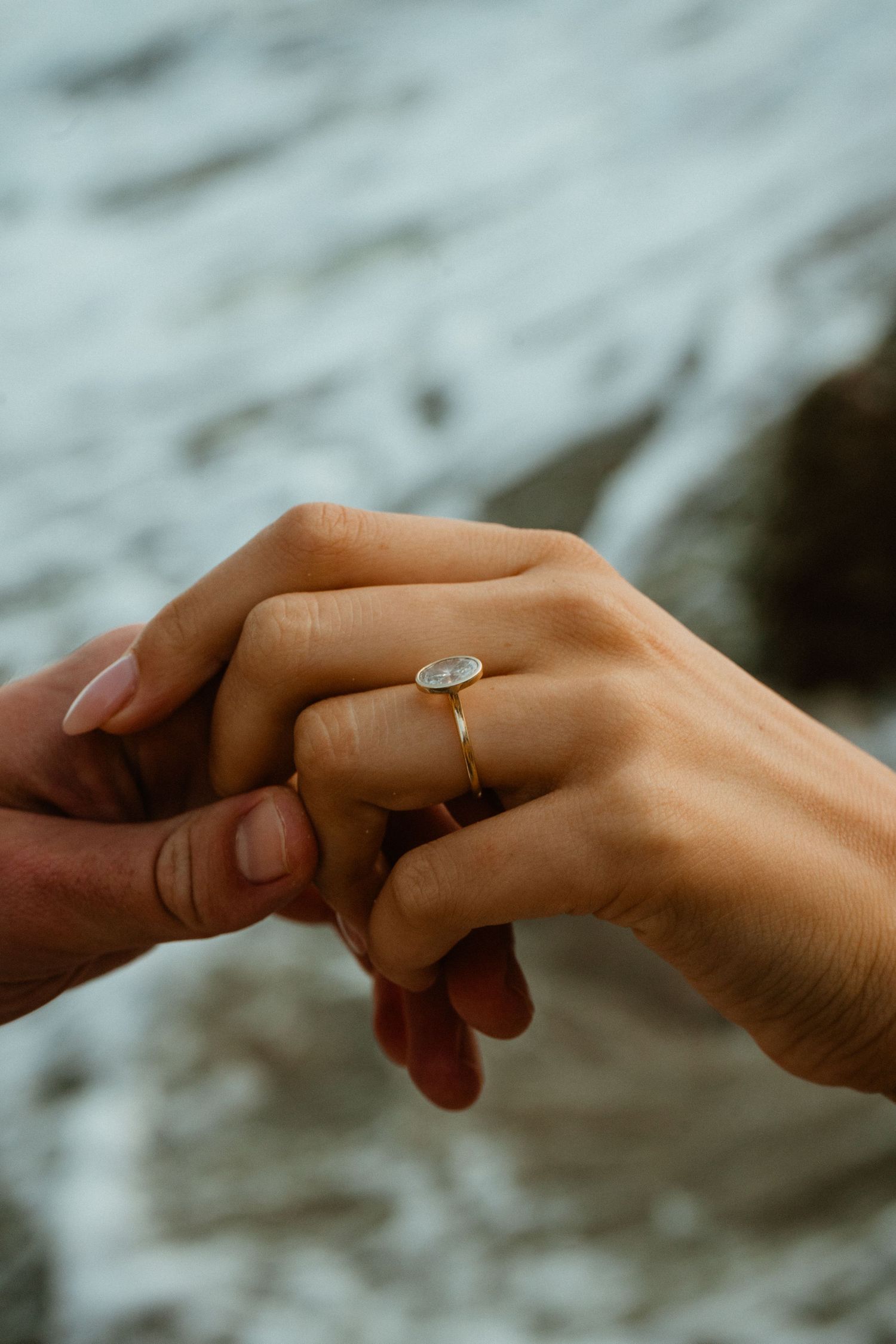 Two hands holding together with an oval diamond engagement ring on a gold band, with rocky ocean waves in the background.