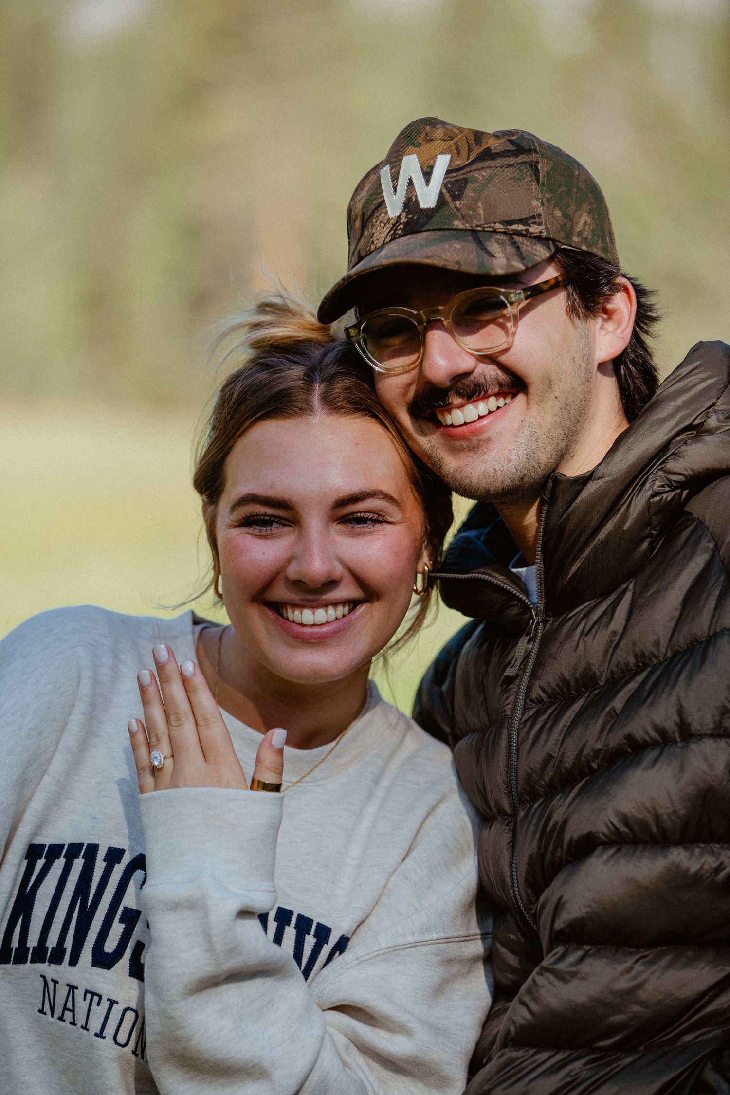 Happy couple smiling after engagement, woman showing off ring, man in camo hat and puffer jacket.
