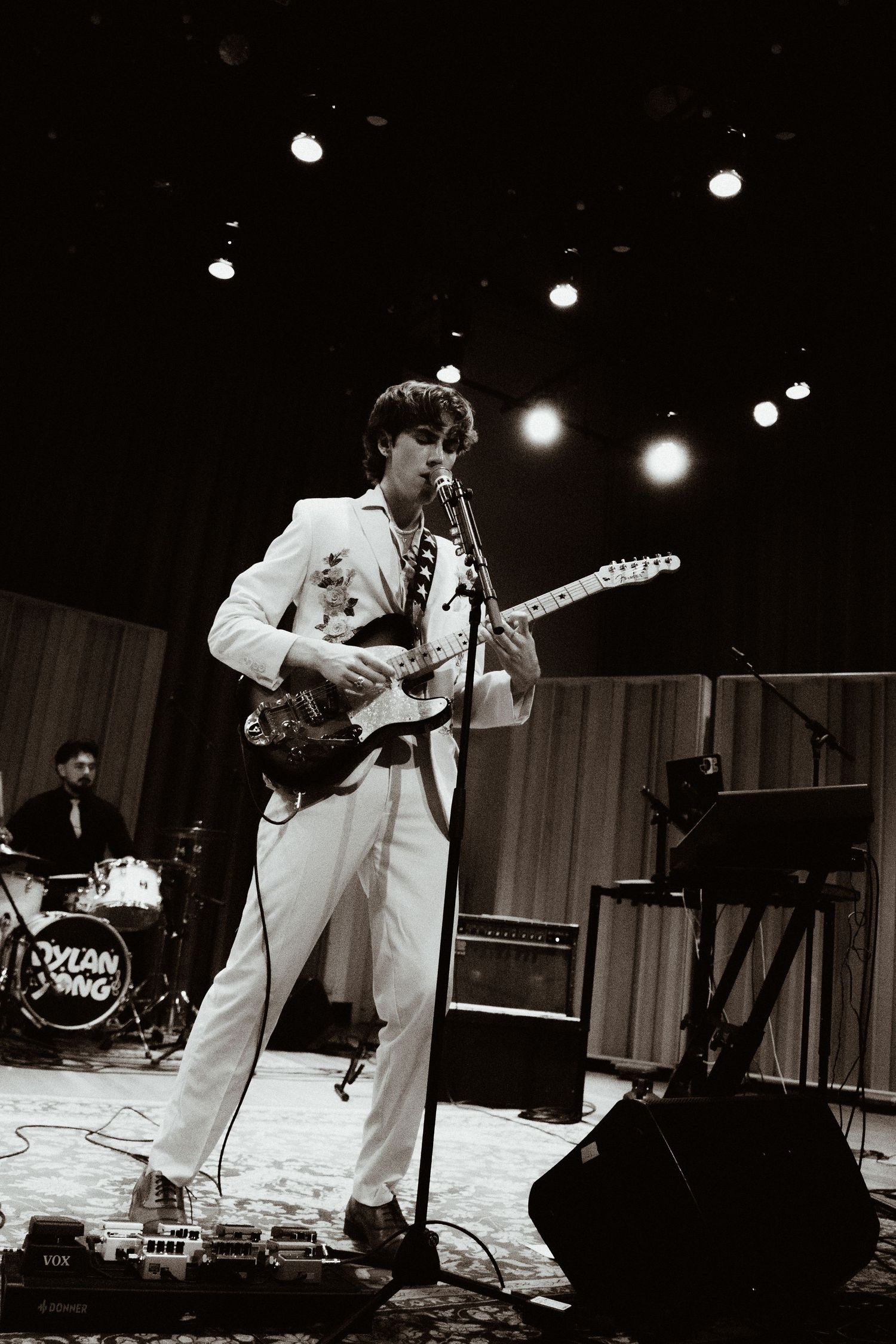 Dylan Yong performing on stage in white suit playing electric guitar in black and white photo.