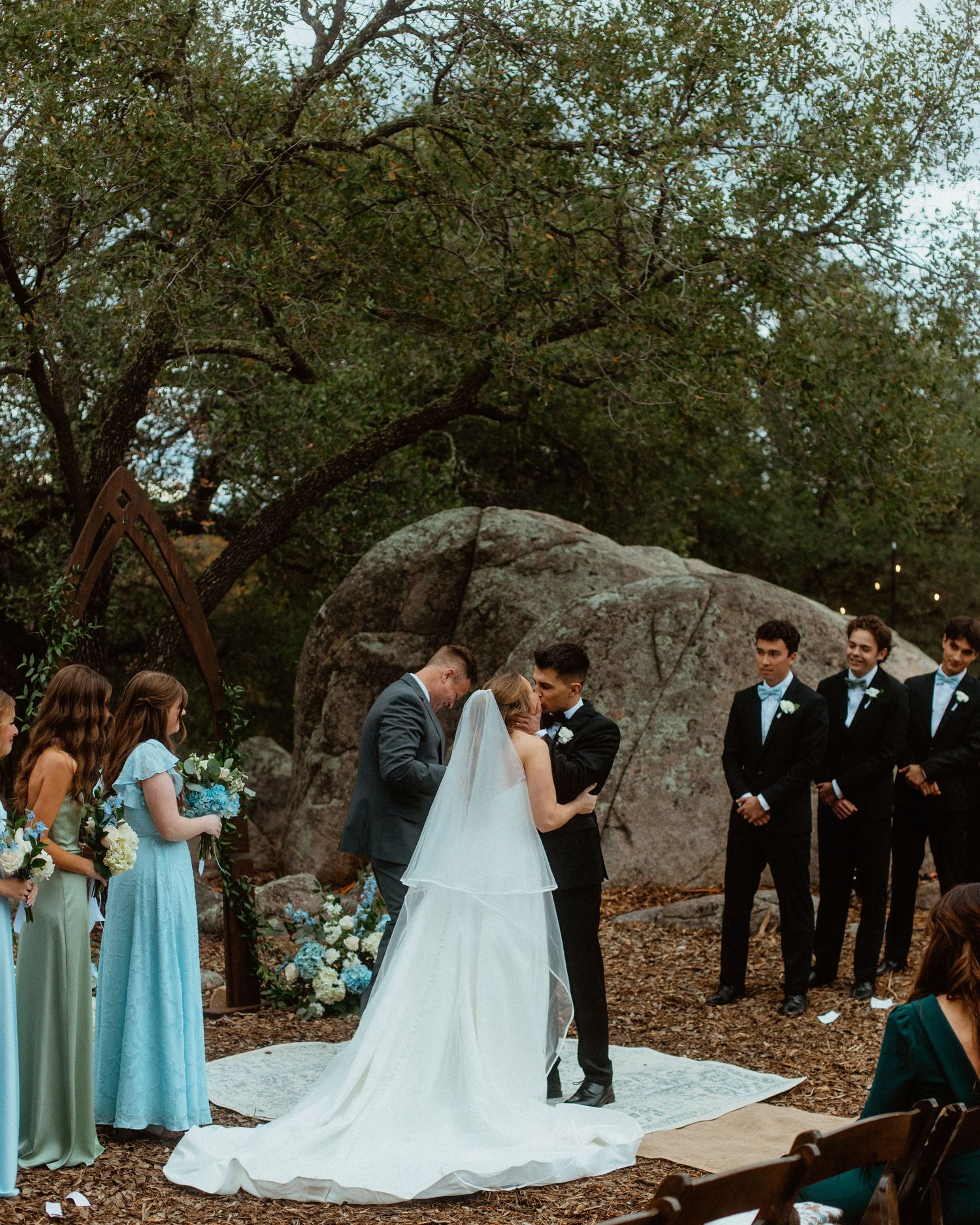 Outdoor wedding ceremony under a large oak tree with bridesmaids and groomsmen in attendance.