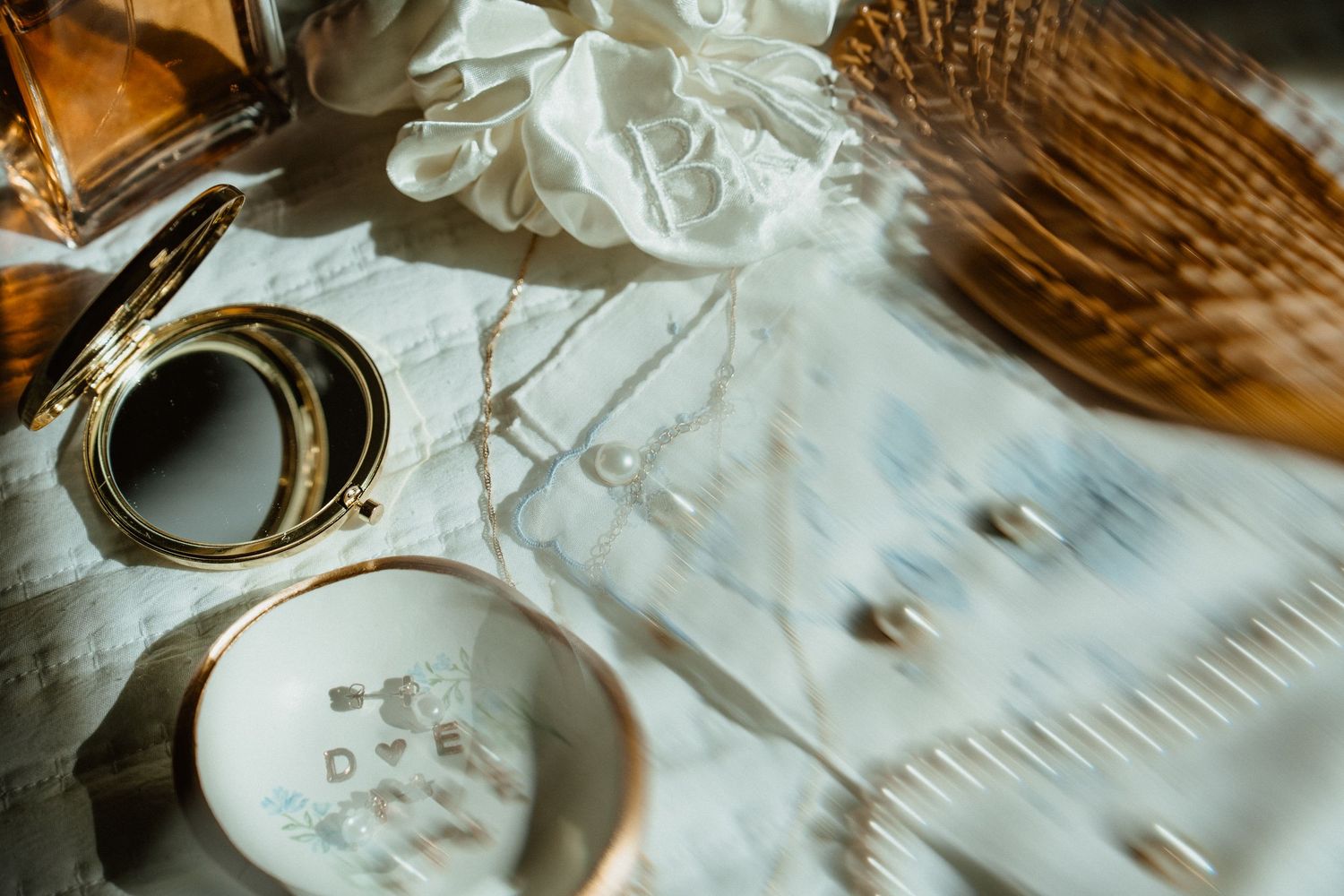 Elegant flat lay of gold jewelry, compact mirror, perfume bottle, and white floral accessories on a marble surface.