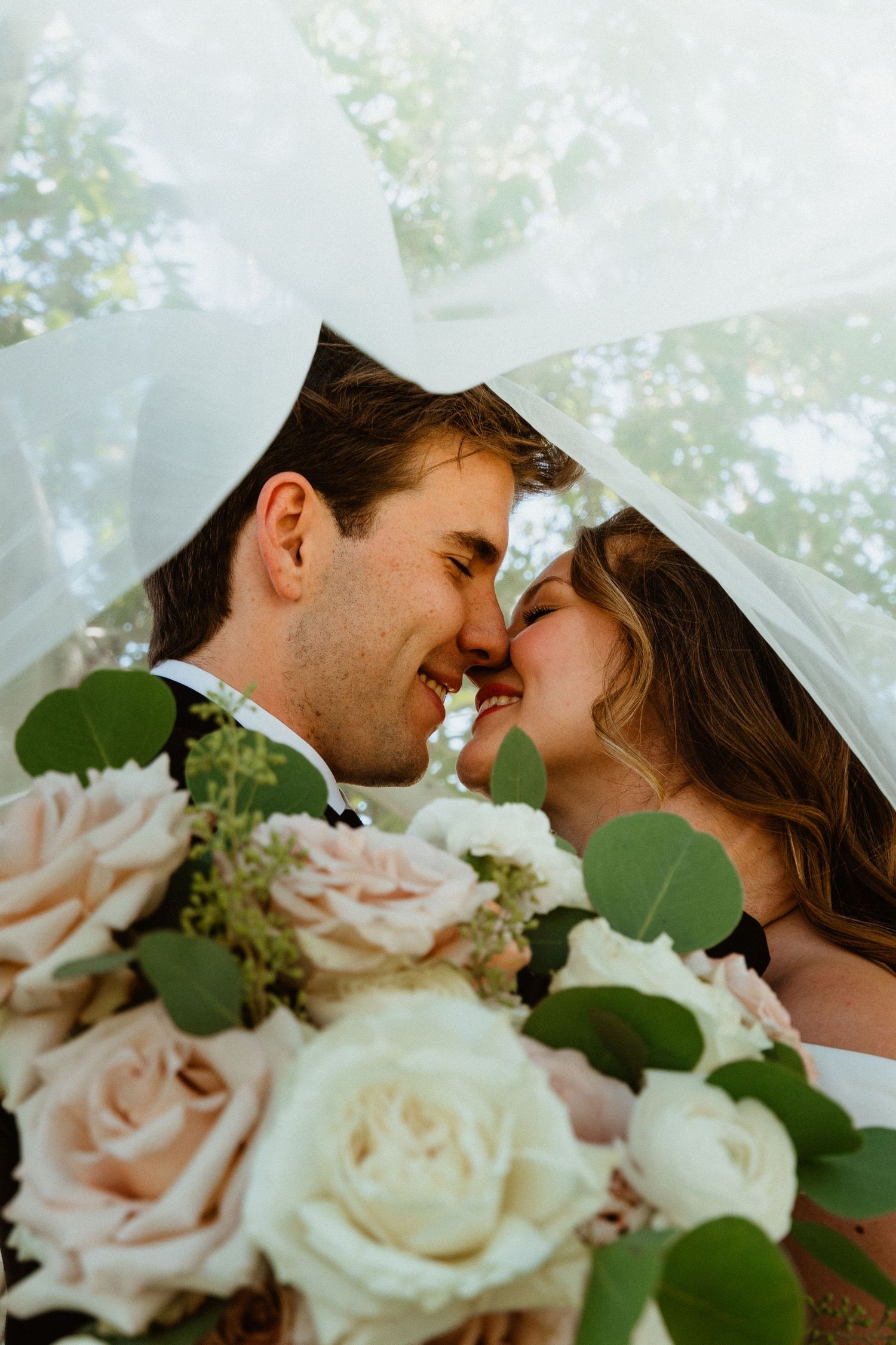 Newlyweds share a romantic nose-to-nose moment under a bridal veil with a white and blush rose bouquet.