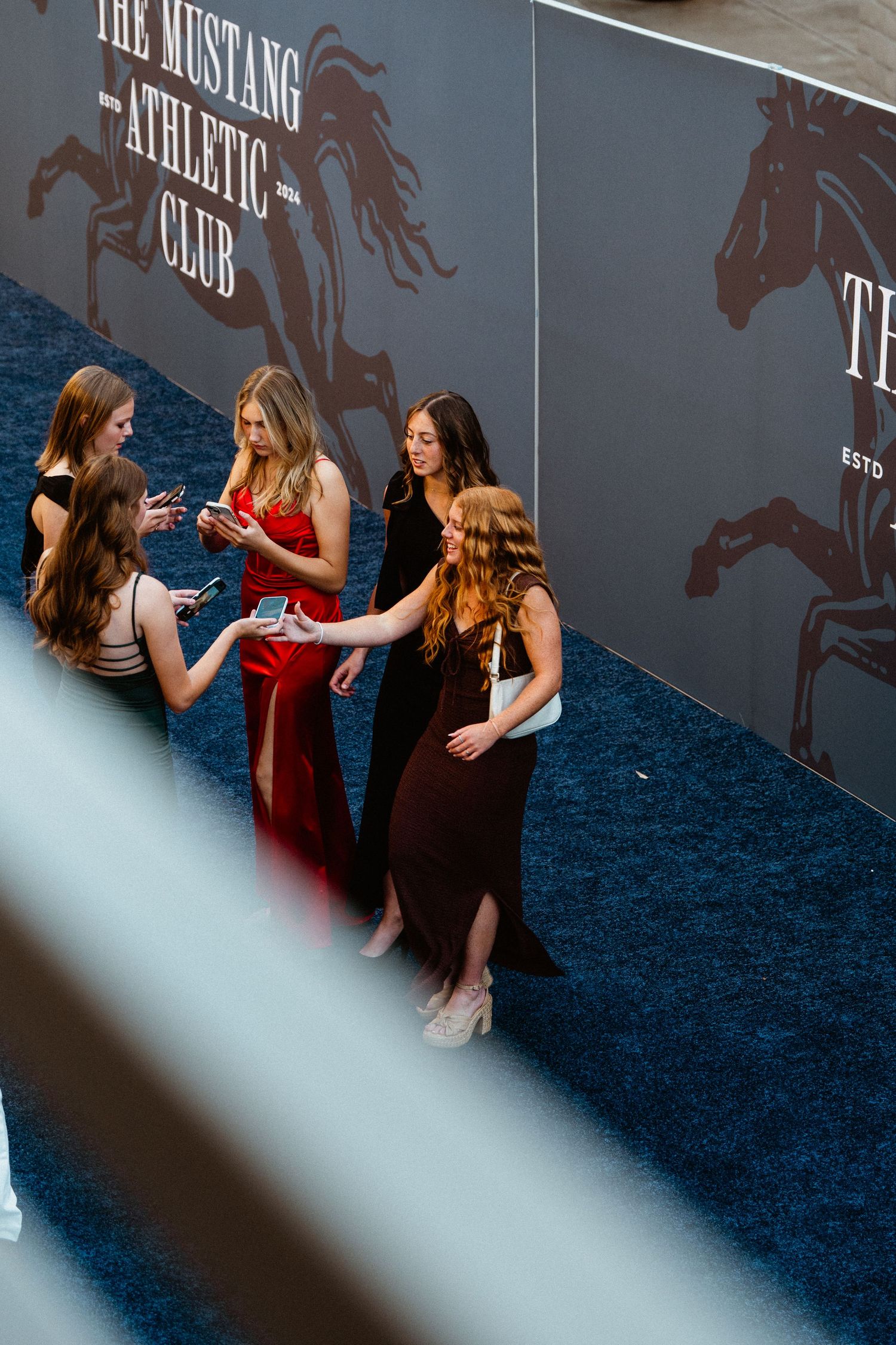 Women in elegant dresses gather on a blue carpet at the Mustang Athletic Club event.