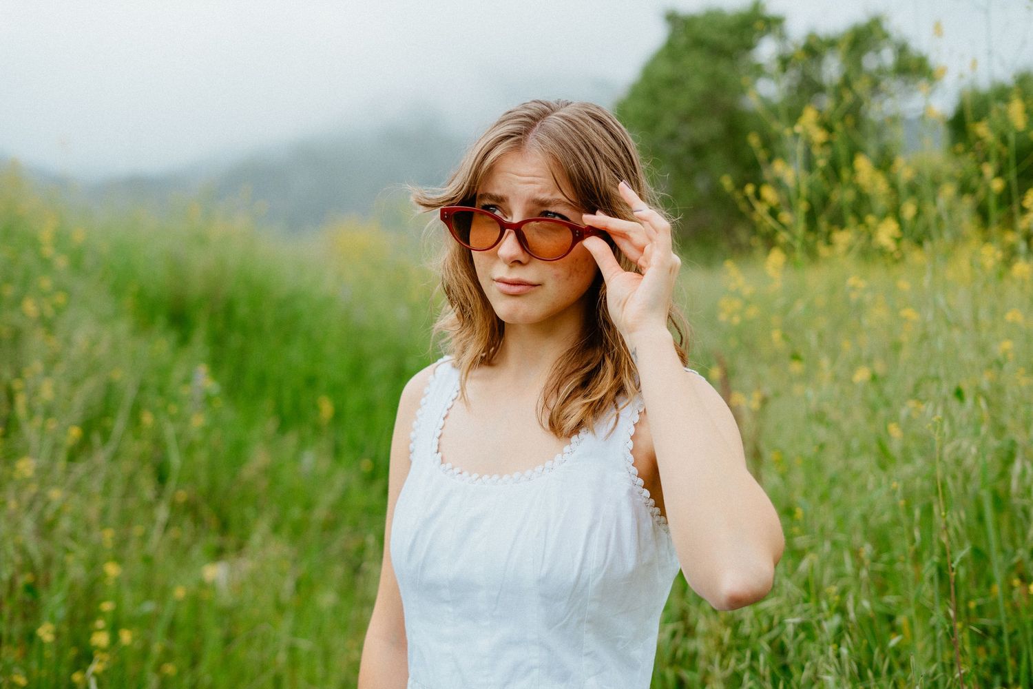 Young woman in white dress gently holds orange sunglasses in a lush green wildflower field.