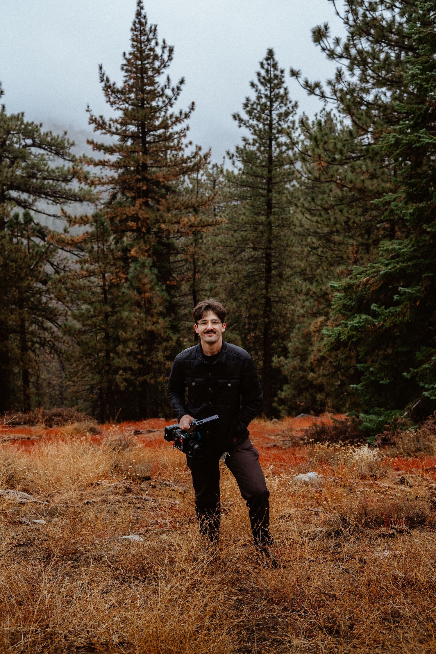 Smiling man holding a camera in an autumn forest clearing with pine trees.