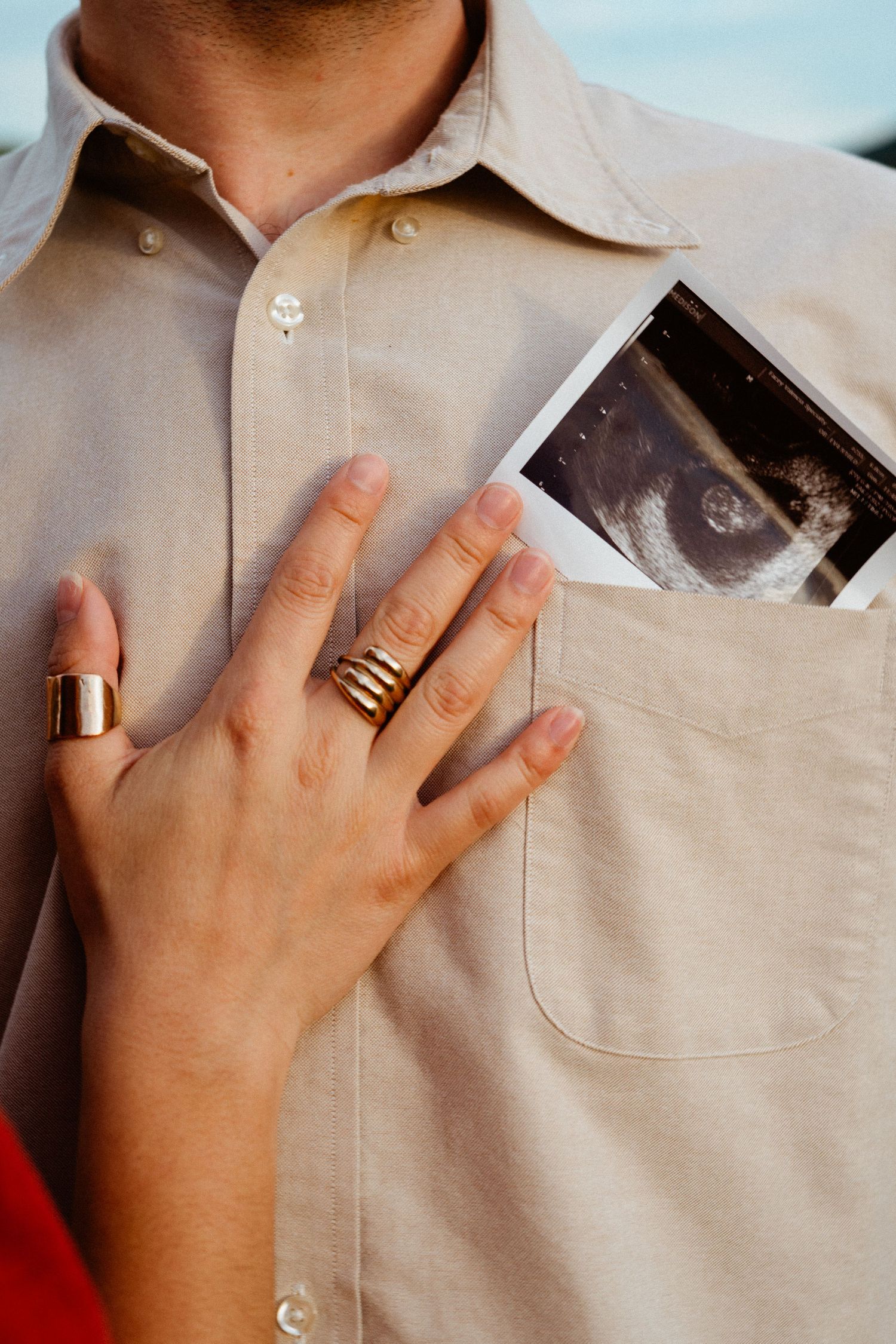 Woman with gold rings places hand on mans chest pocket holding ultrasound photo announcement.