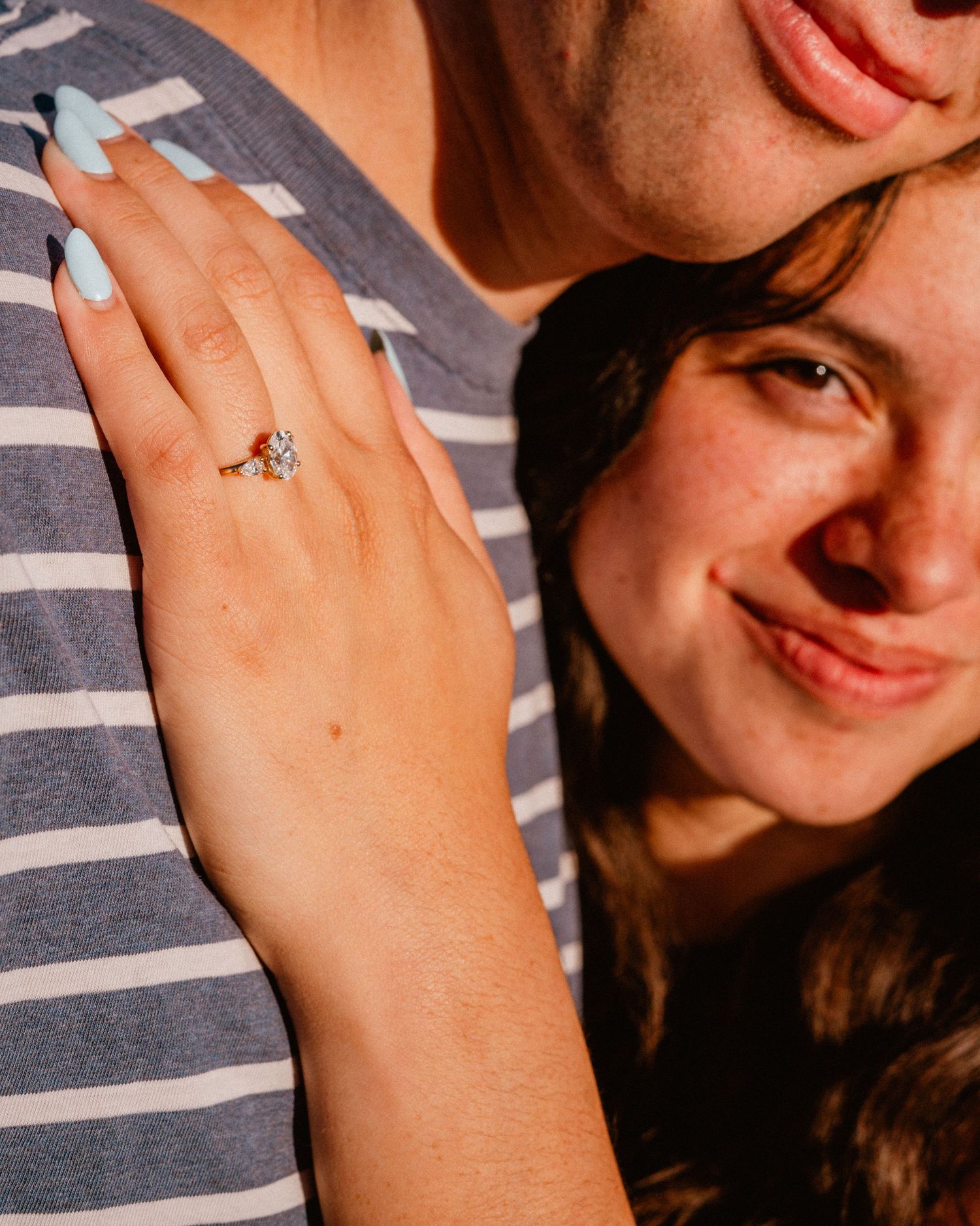 Smiling woman showing off her diamond engagement ring on her partners striped shirt in warm golden light.