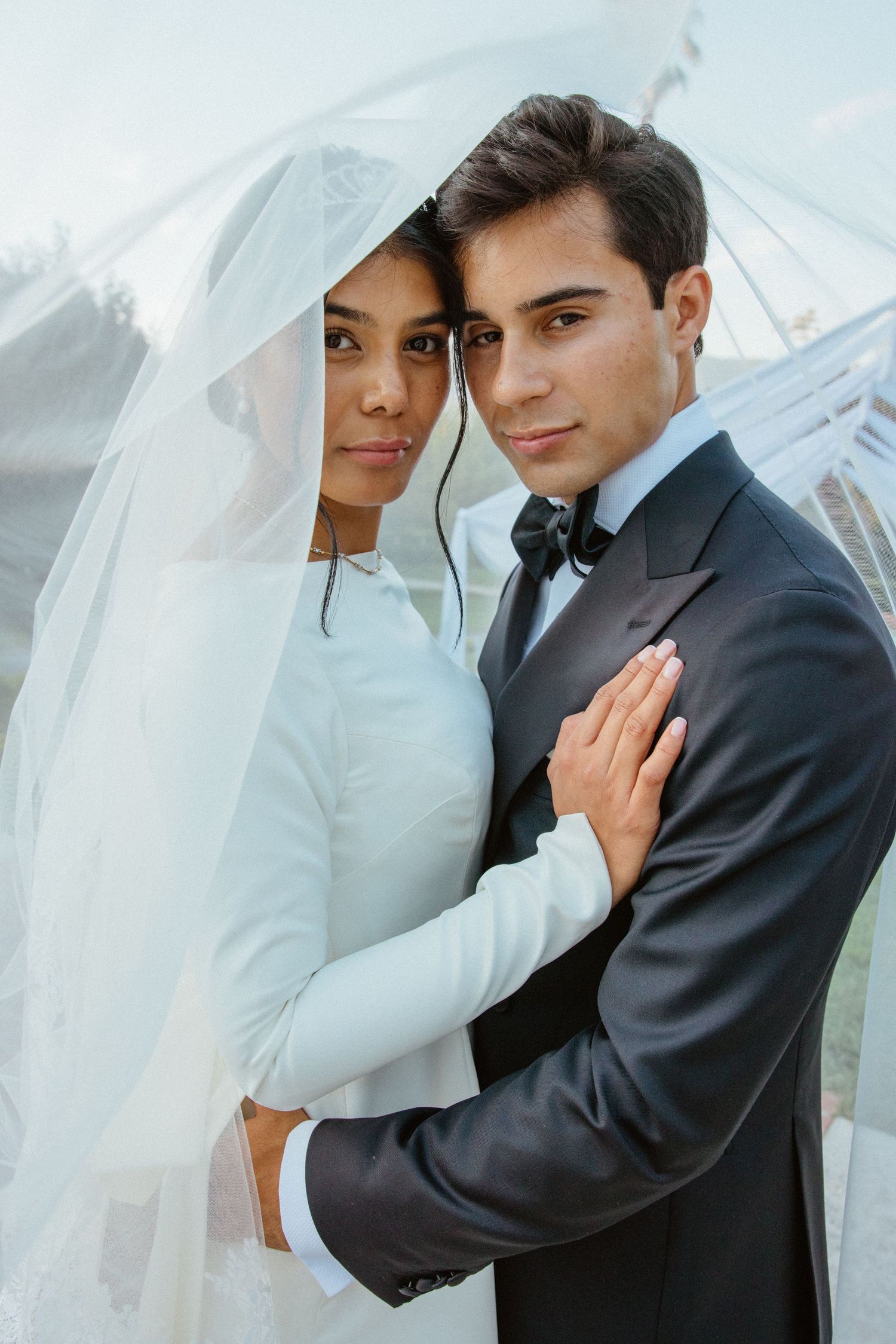 Bride and groom embrace under flowing white veil, posing together at outdoor wedding ceremony venue.