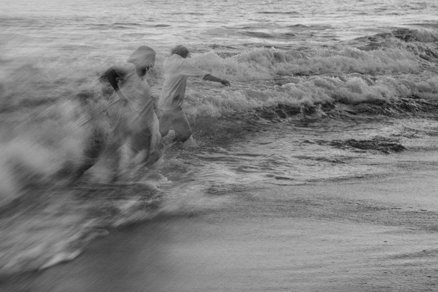 Blurry black and white photo of people running into ocean waves on a beach, captured with motion blur effect.